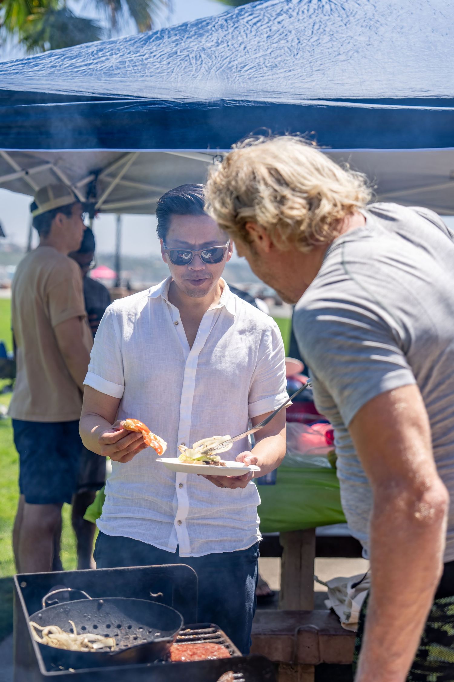 Asian man in white shirt receiving grilled food on a plate at an outdoor beach BBQ event.