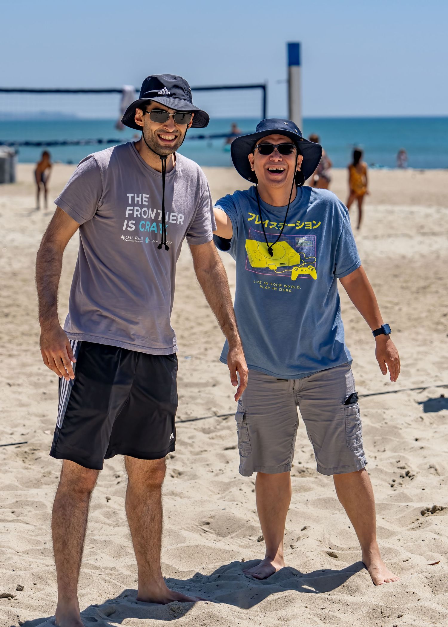 Two smiling men in bucket hats standing together on a sunny beach volleyball court.