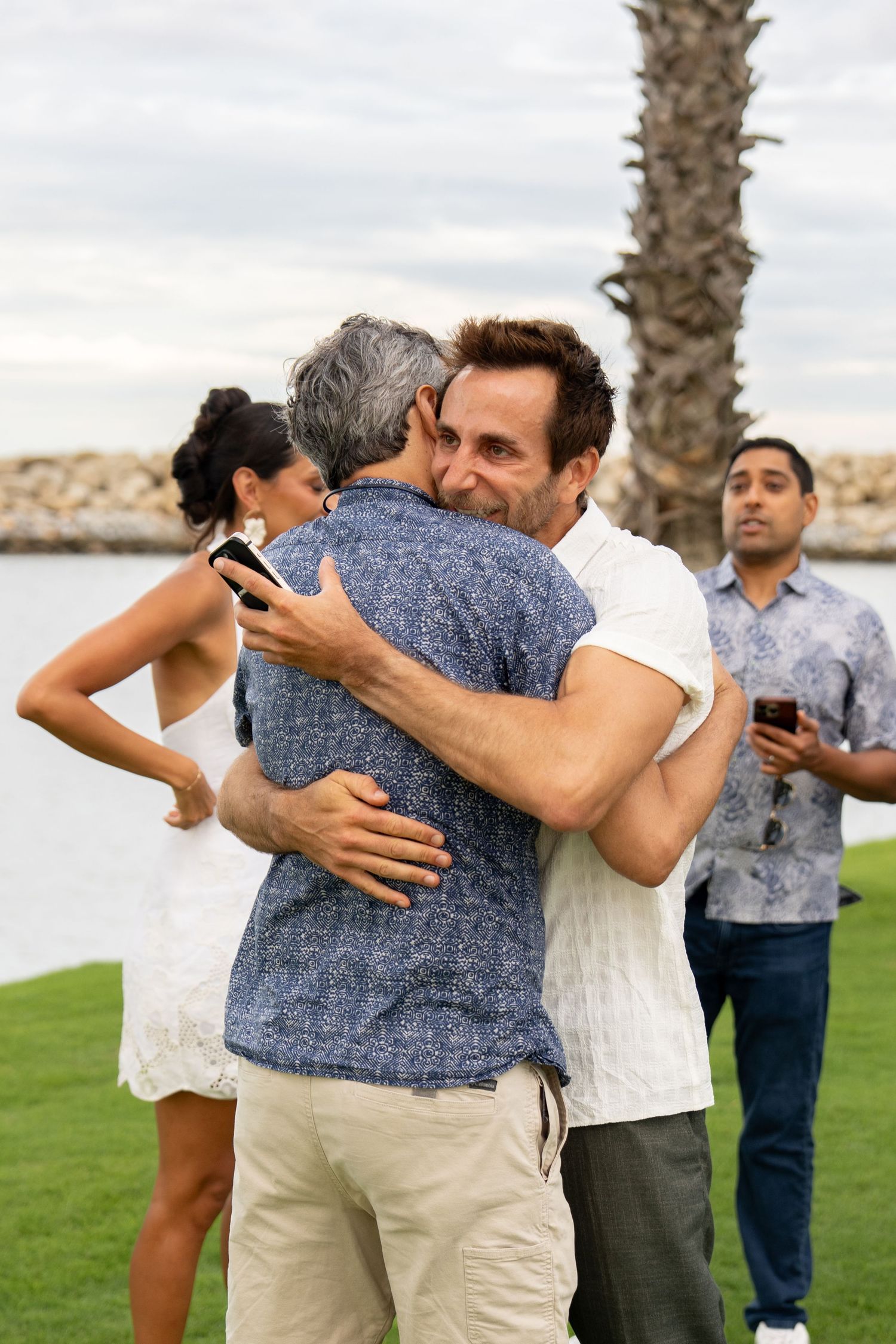 Two men hugging warmly at an outdoor waterfront gathering with palm trees.