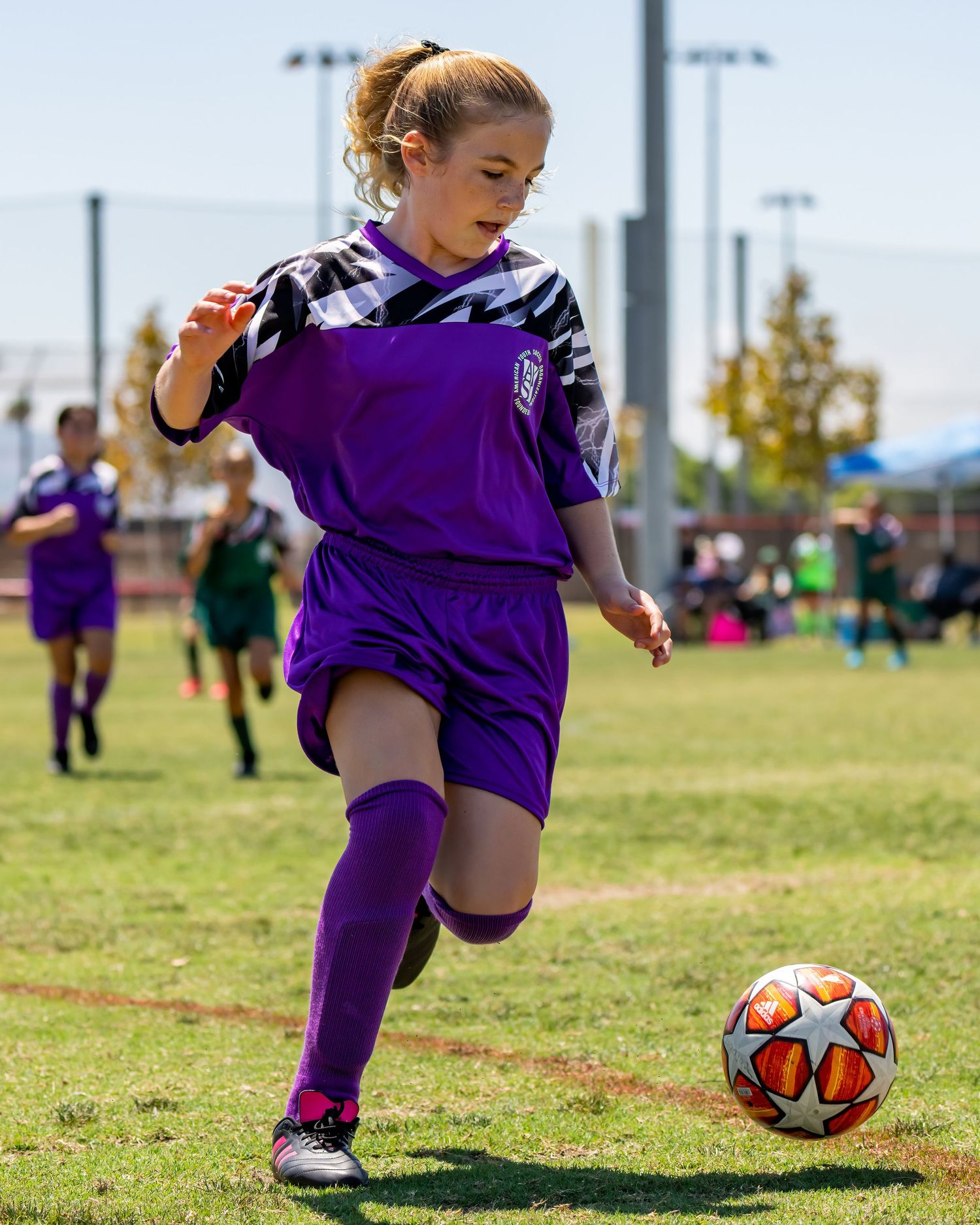 Young girl in purple soccer kit dribbling white soccer ball during outdoor youth match.