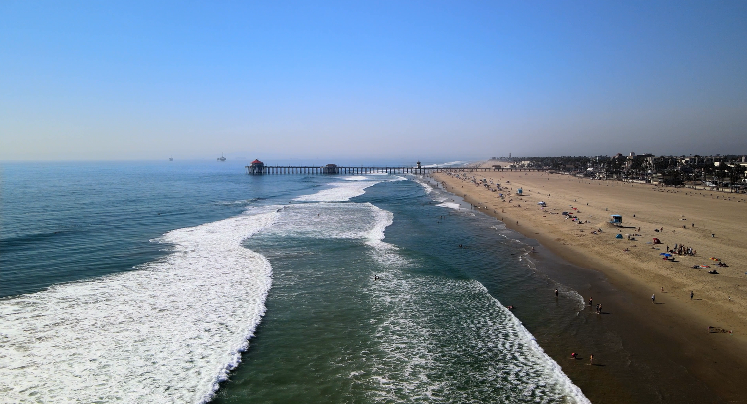 Aerial view of a sandy beach with rolling ocean waves under a clear blue sky.