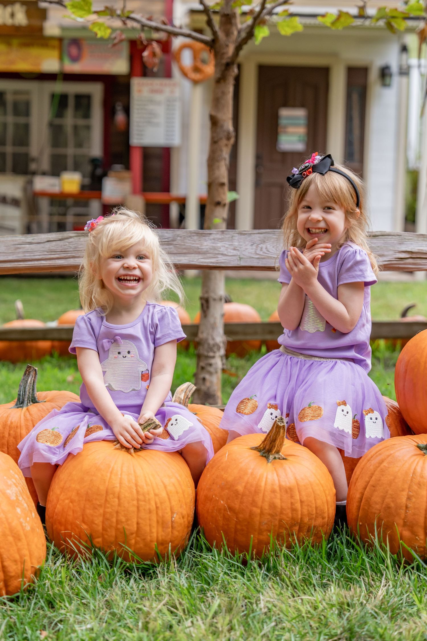 Two blonde sisters in matching purple Halloween outfits sitting on large orange pumpkins.