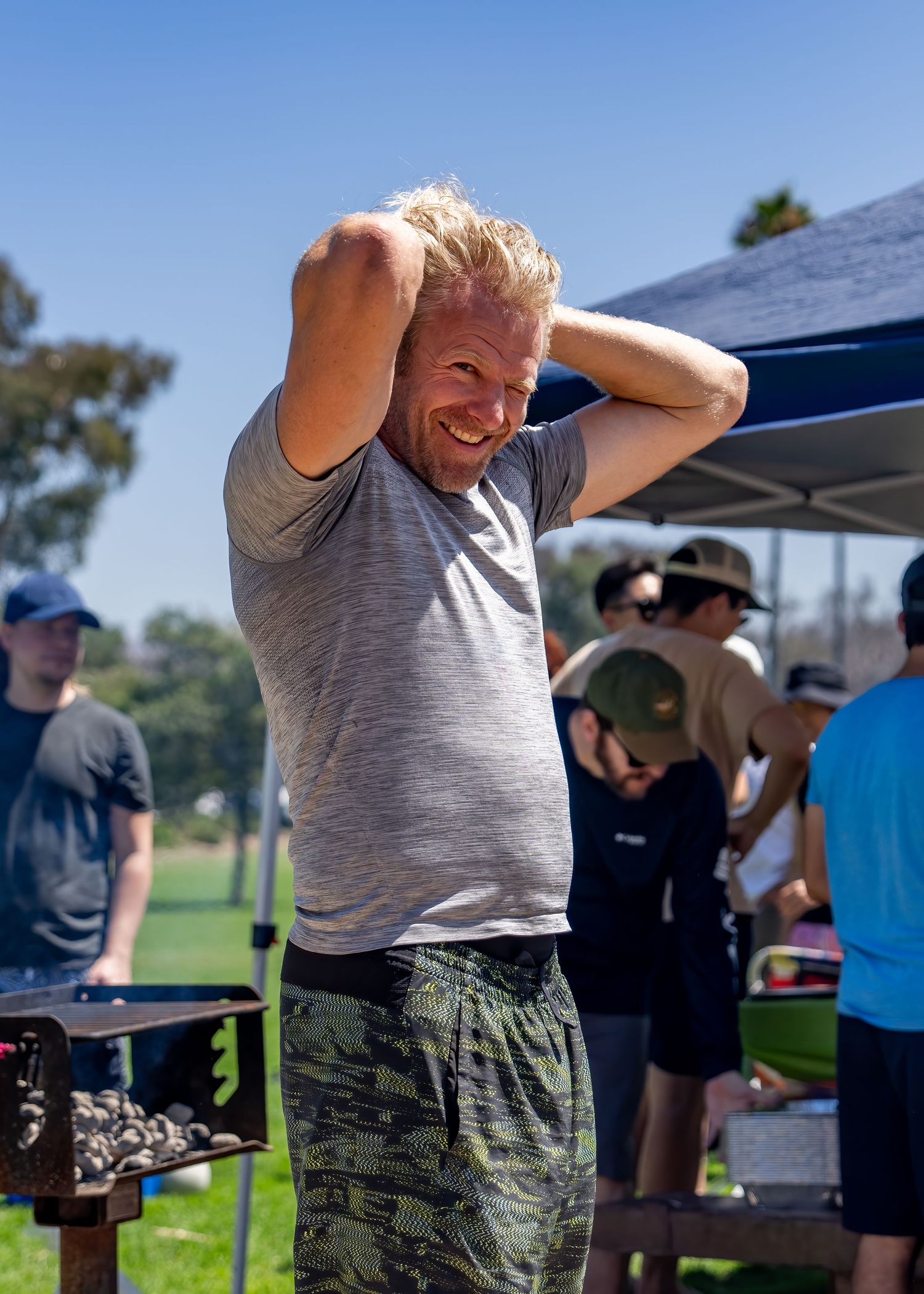 Laughing blond man with hands behind his head standing near a grill at an outdoor park event.