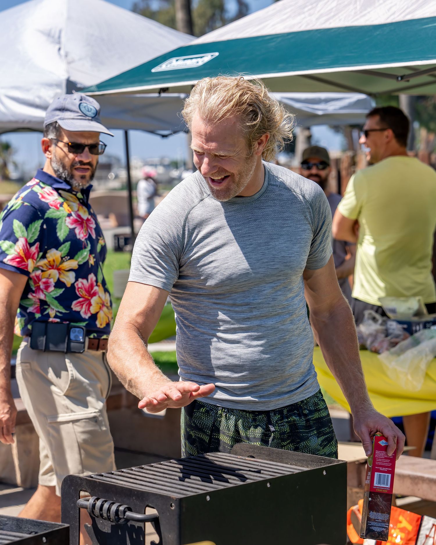 Smiling blond man grilling outdoors at a sunny park event with canopy tents.