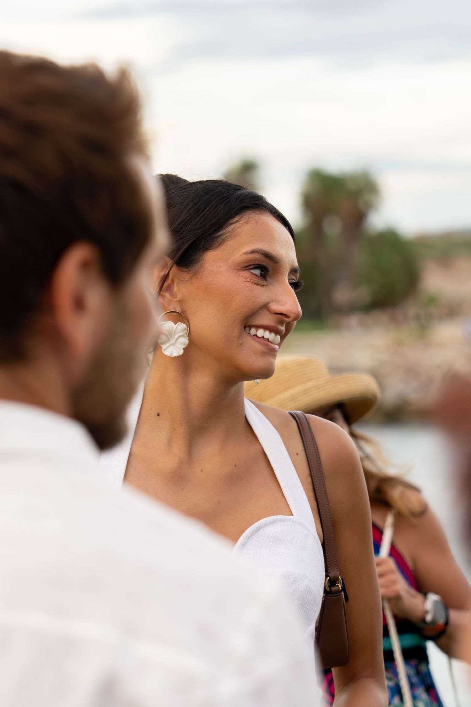 Smiling woman in white halter dress with large floral earrings at outdoor event.