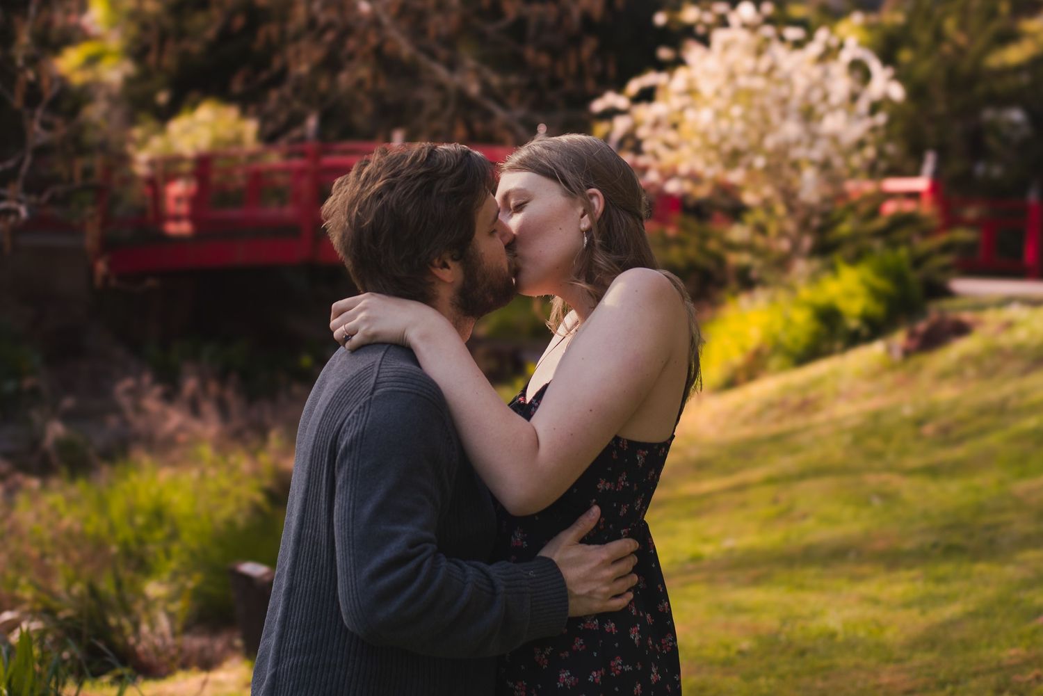 A photo of a couple kissing in front of a bridge.