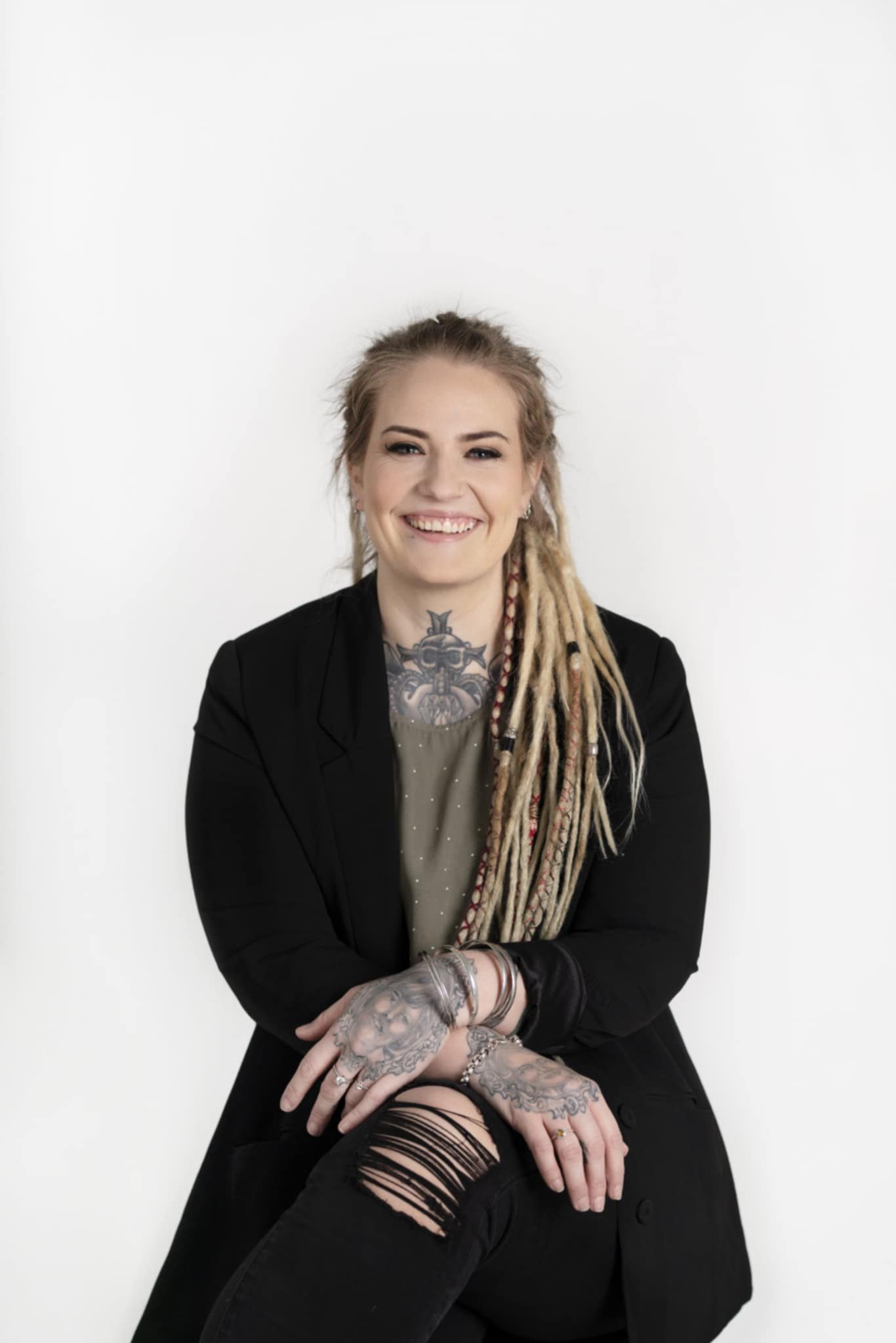 Smiling woman with blonde dreadlocks, tattoos on chest and hands, wearing black blazer and ripped jeans, seated against white background.