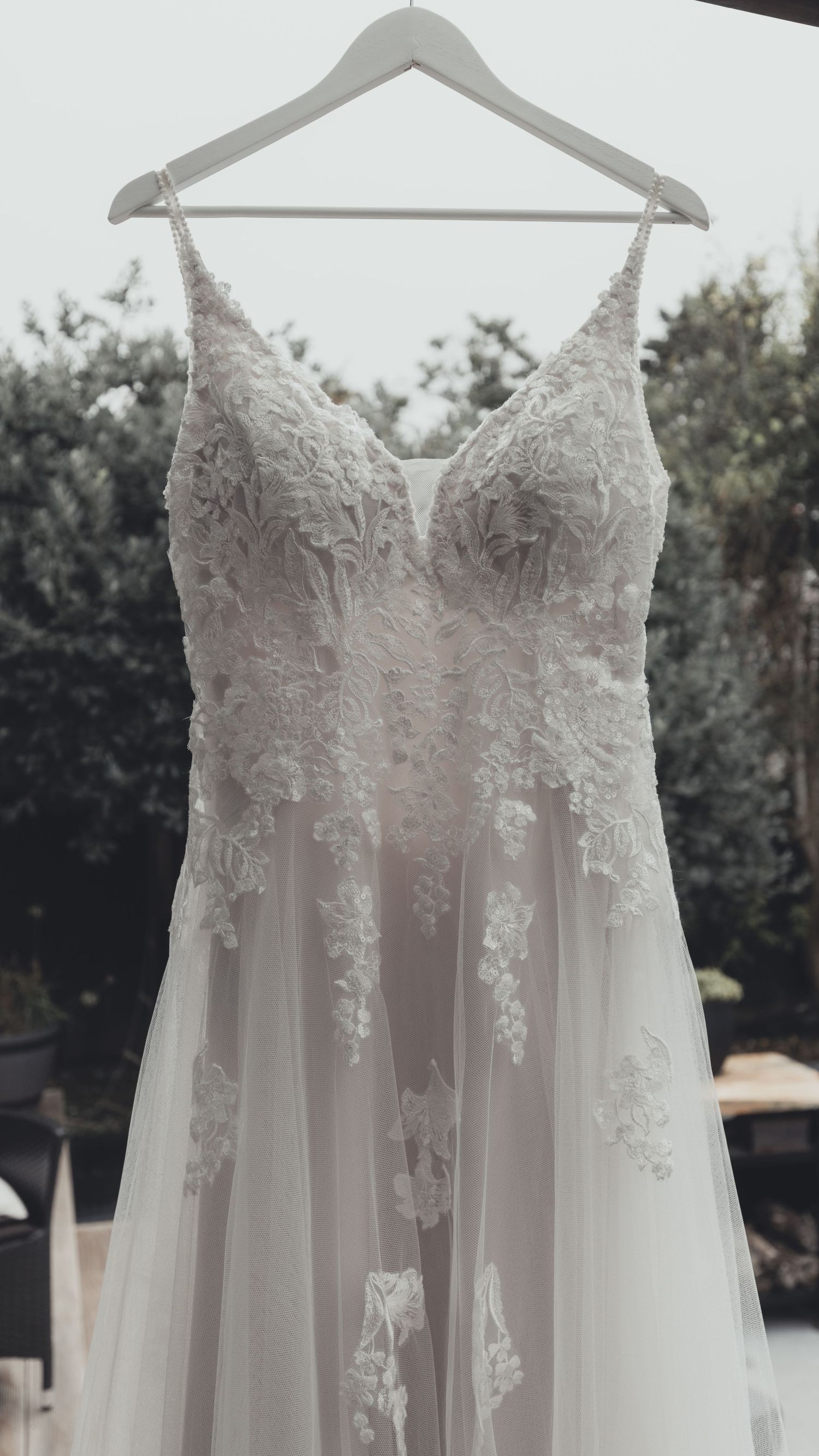 An elegant lace and tulle wedding dress hangs on a hanger outdoors.