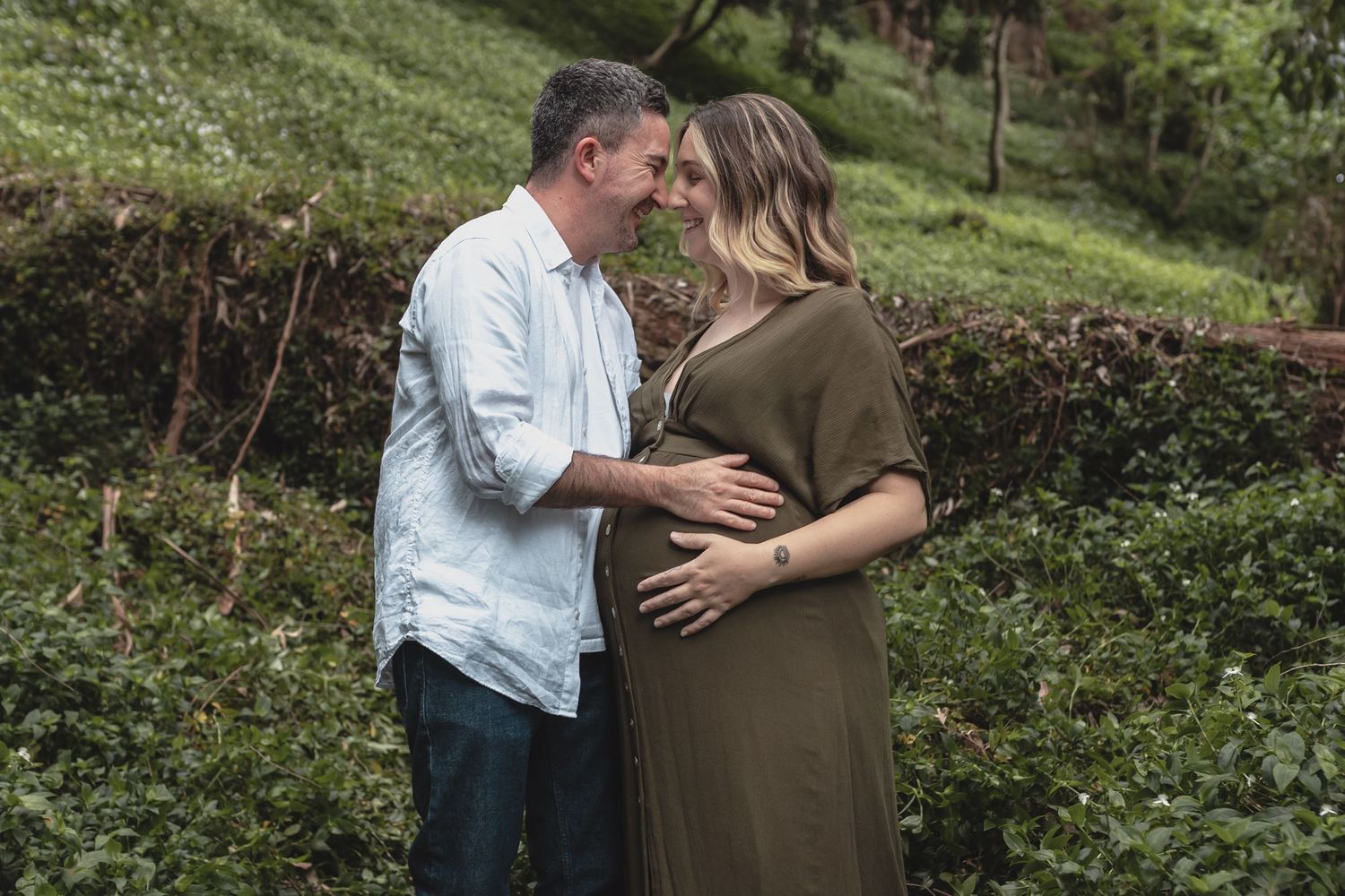 Expectant couple sharing an intimate forehead touch in a lush green outdoor setting.