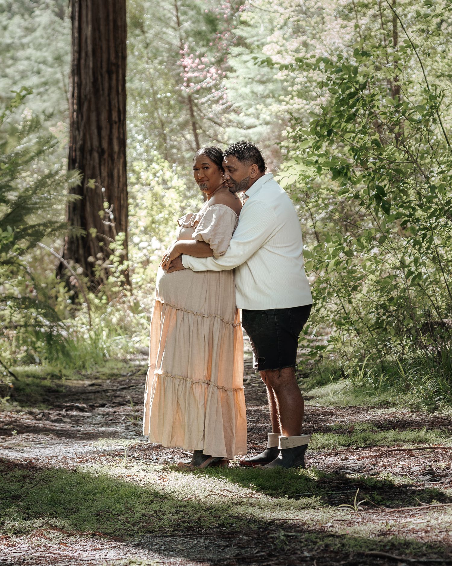 Couple embracing in a sunlit forest, woman in a flowy peach maternity dress, man in white shirt and black shorts.