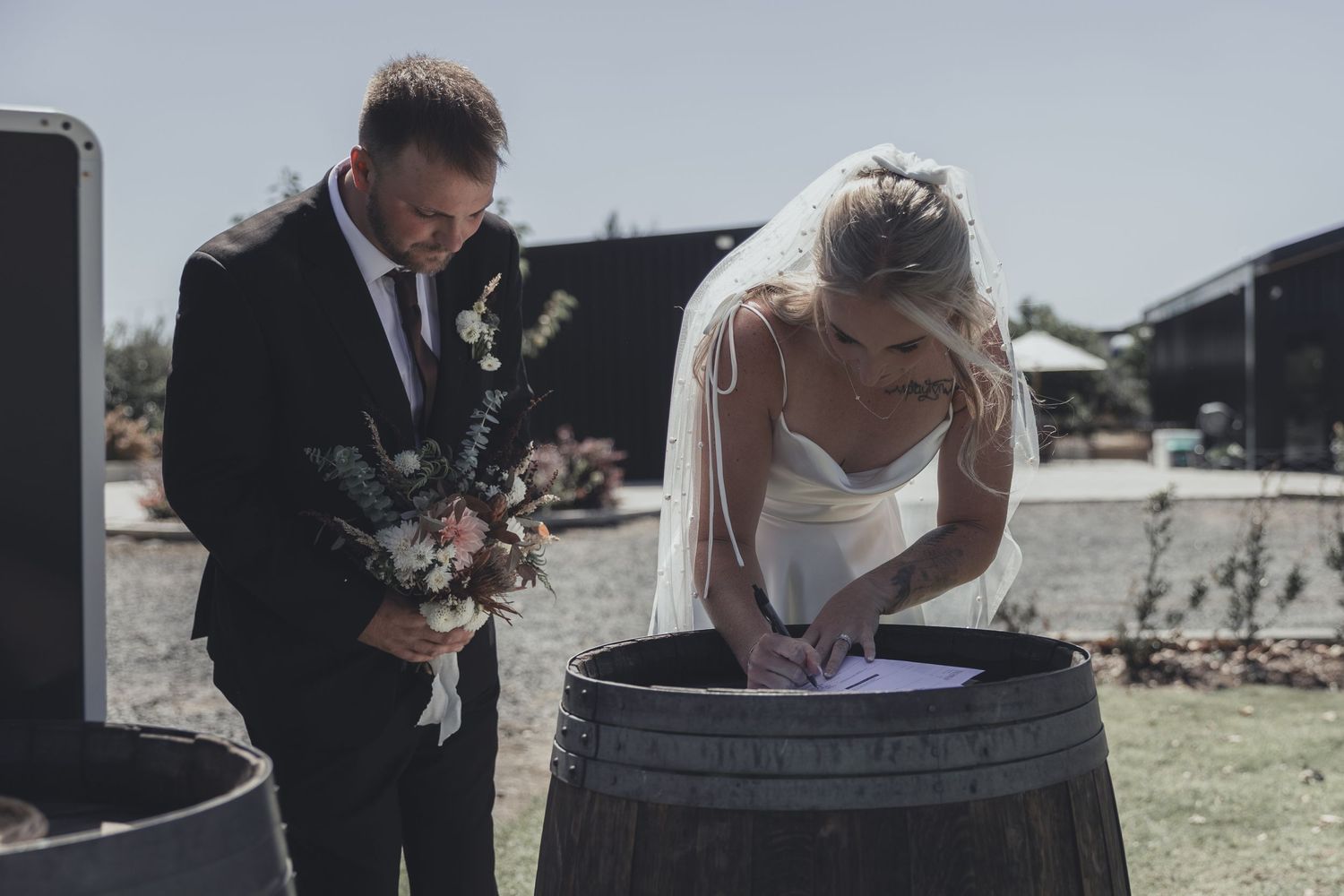 Bride signs wedding documents on a wooden barrel while groom holds bouquet at outdoor rustic venue.