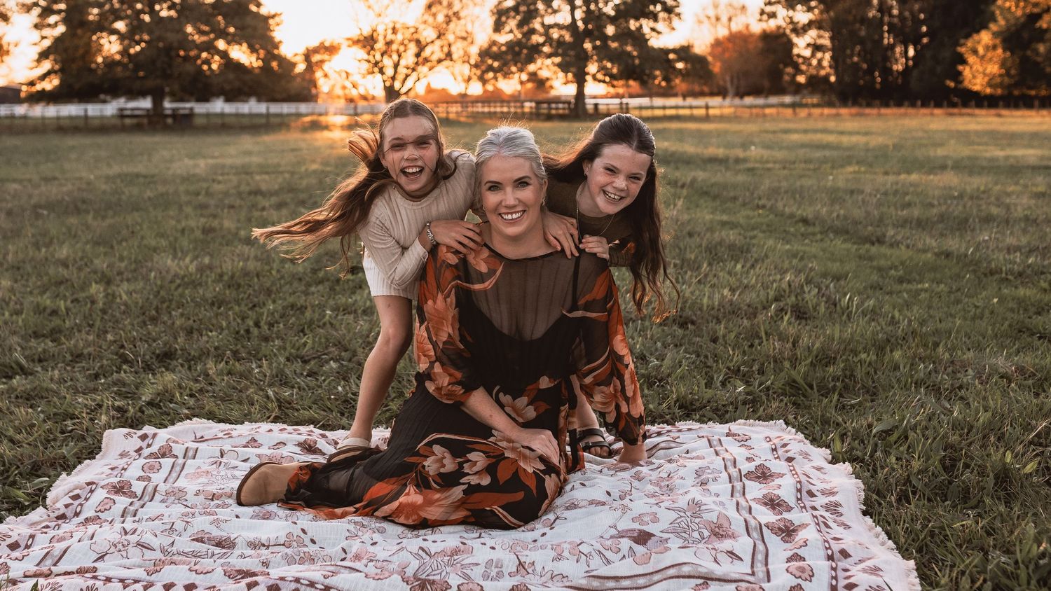 Happy mother sitting on blanket with two laughing daughters during golden sunset family photo session.