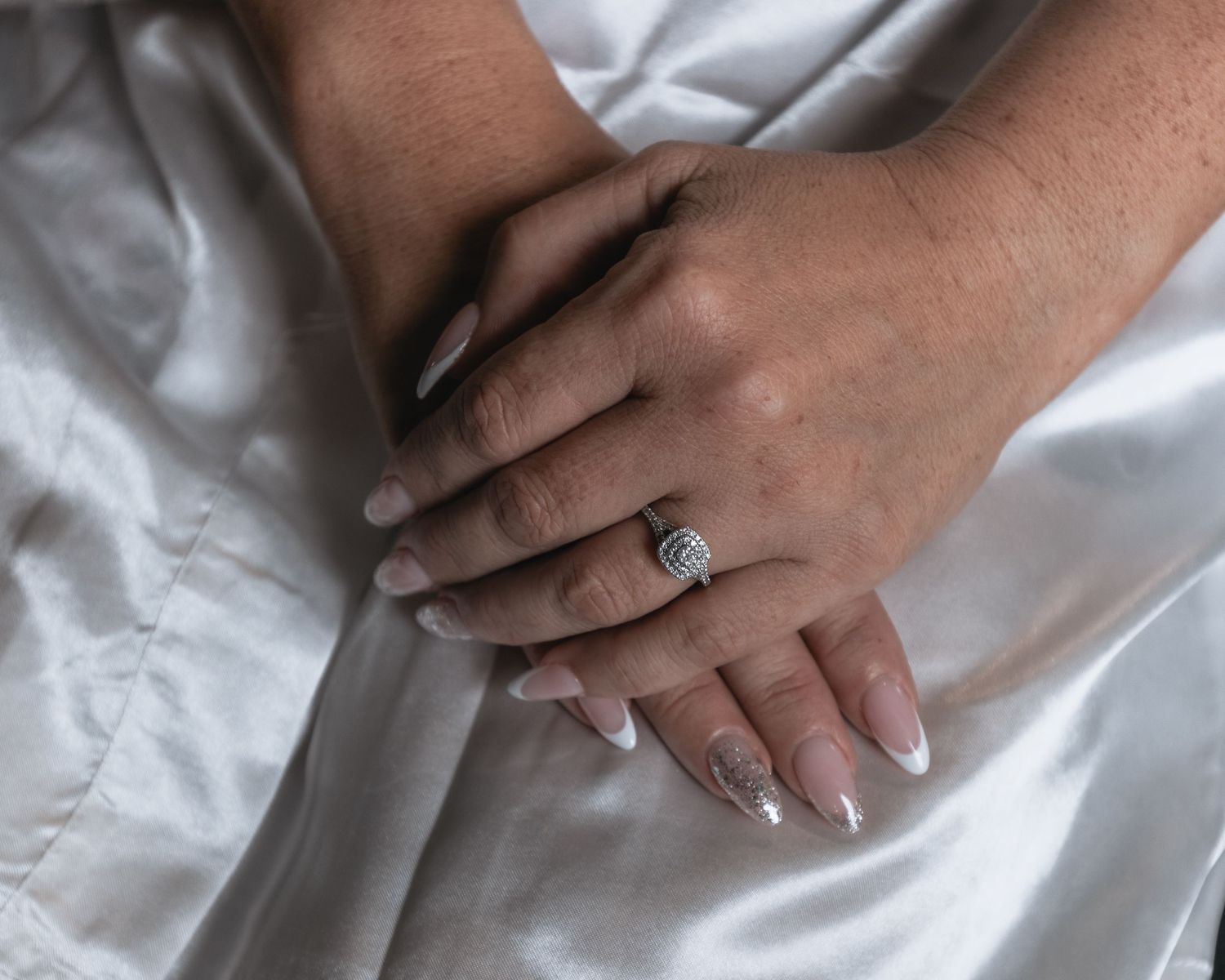 A bride with manicured nails displays her diamond engagement ring on white fabric.