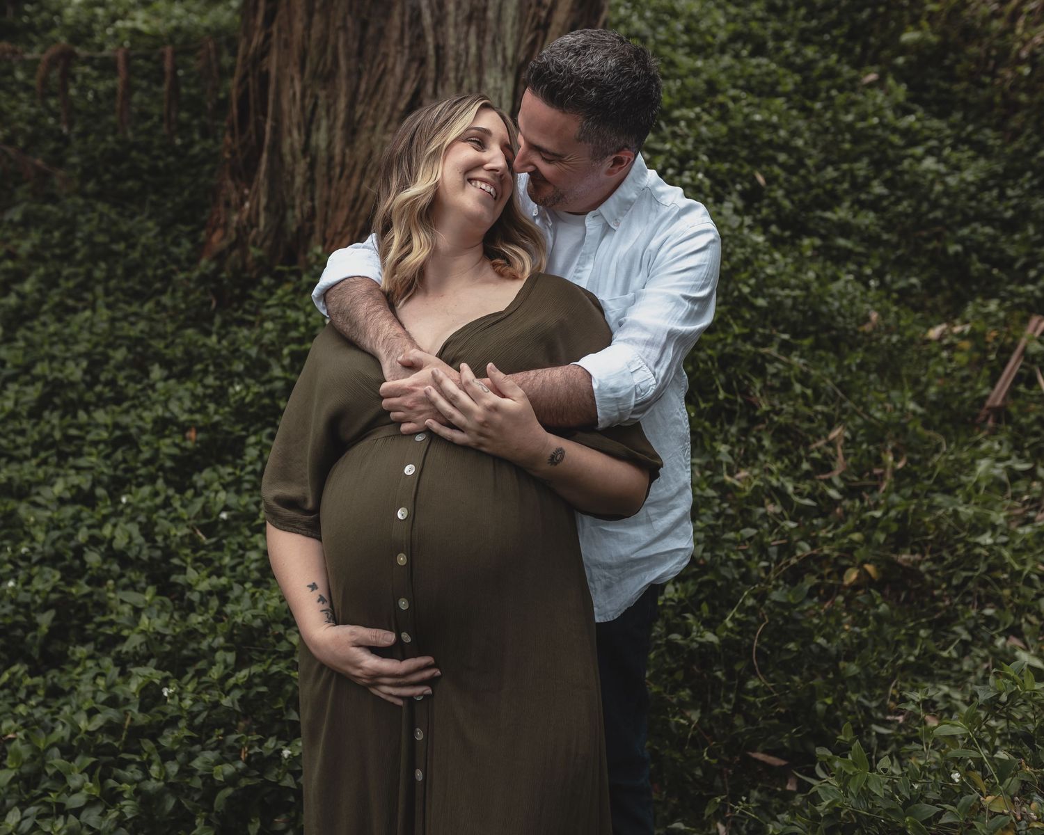 Smiling couple embracing outdoors, pregnant woman in olive dress cradling baby bump near greenery.
