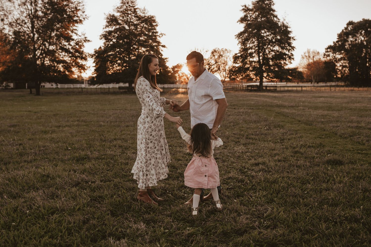 Family of three holding hands in a sunlit field at sunset during outdoor photoshoot.