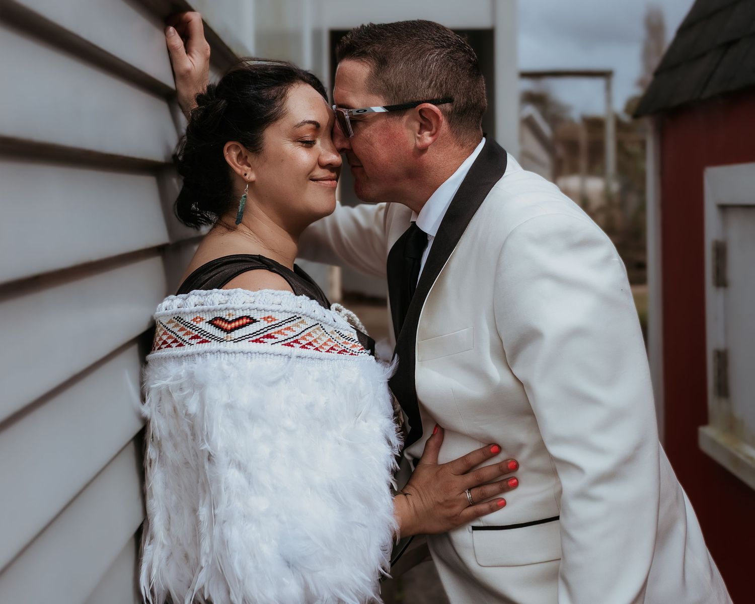 A couple shares a tender kiss outdoors, the woman wearing a traditional Maori korowai cloak at their wedding.