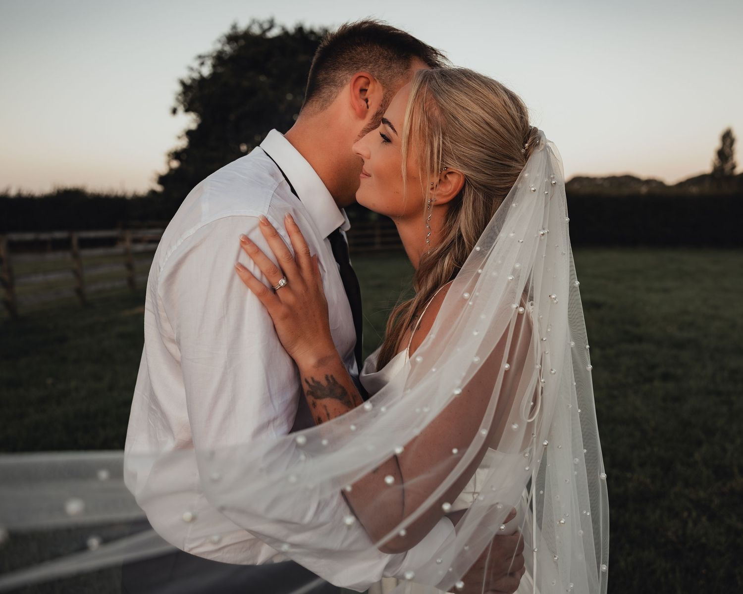 Couple sharing a tender moment at sunset, bride in white gown with pearl veil, groom in white shirt.