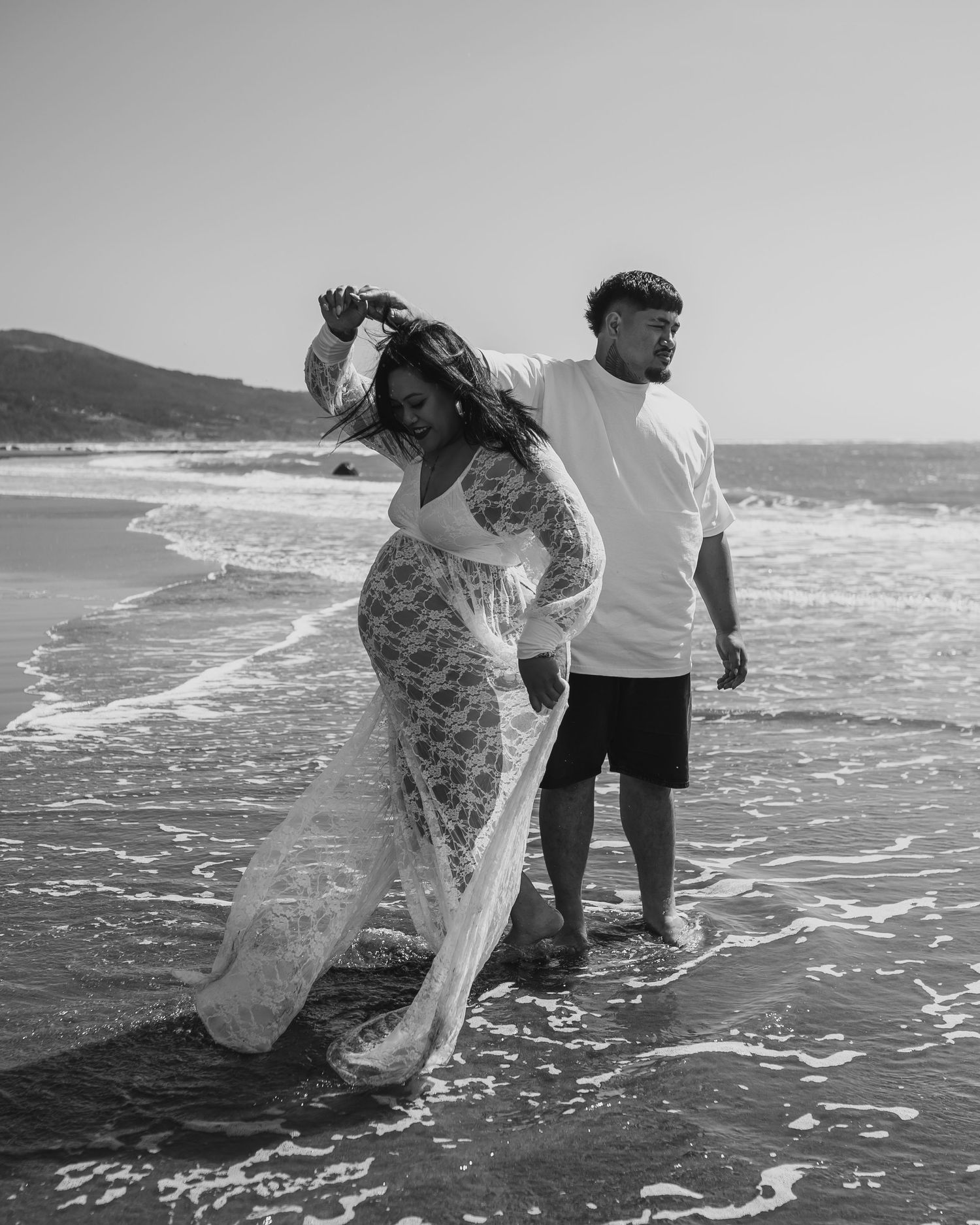 Black and white maternity photo of a couple standing in ocean waves, woman in lace gown holding her hair.