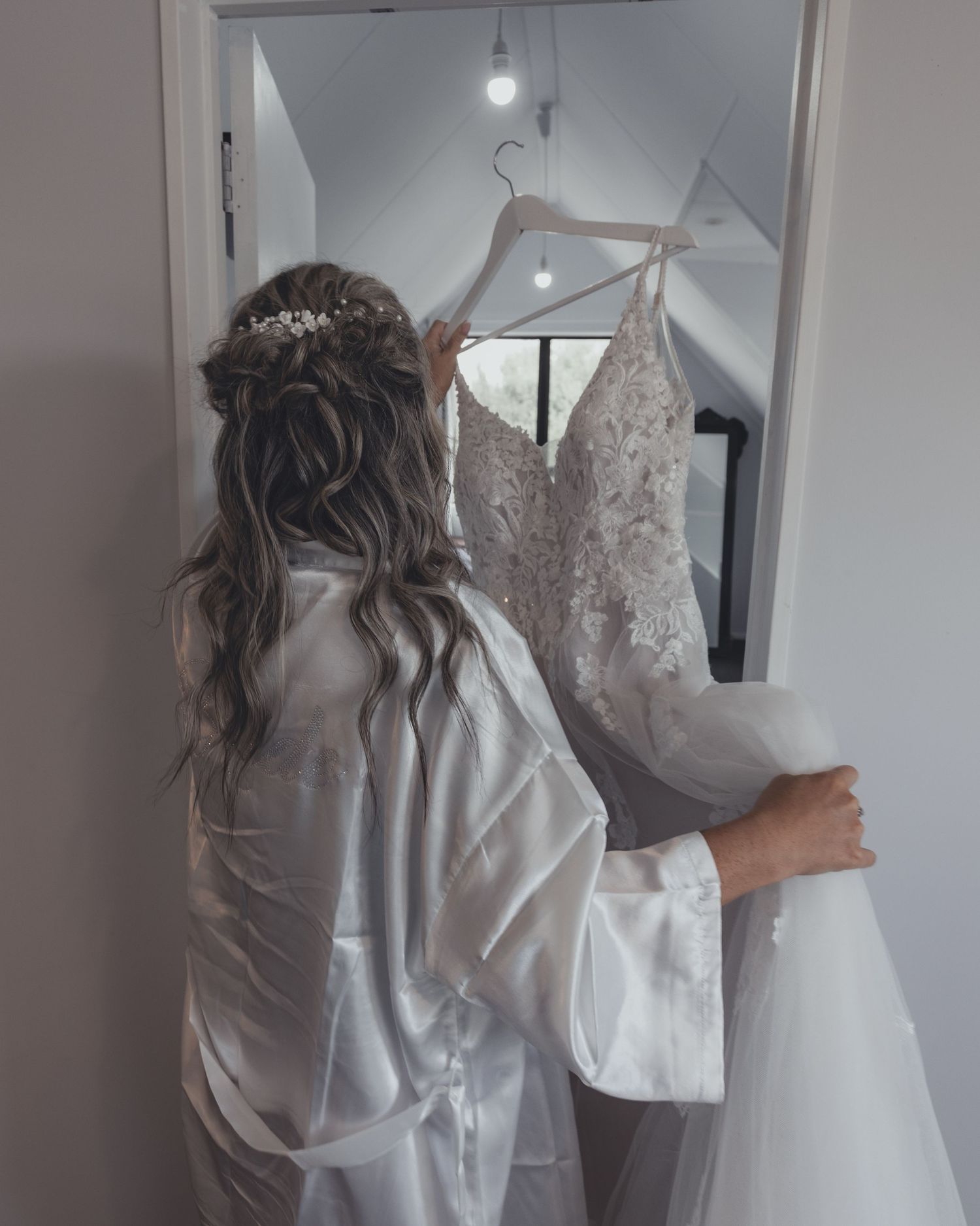 Bride in white robe reaching for lace wedding dress on hanger in bridal suite.