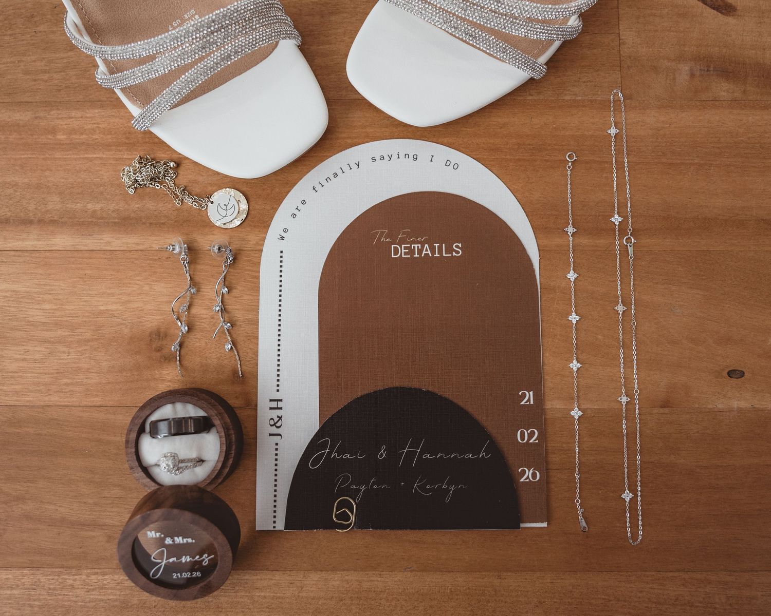 Elegant wedding flat lay with white heels, acrylic details card, ring box, earrings, and delicate jewelry on wood.