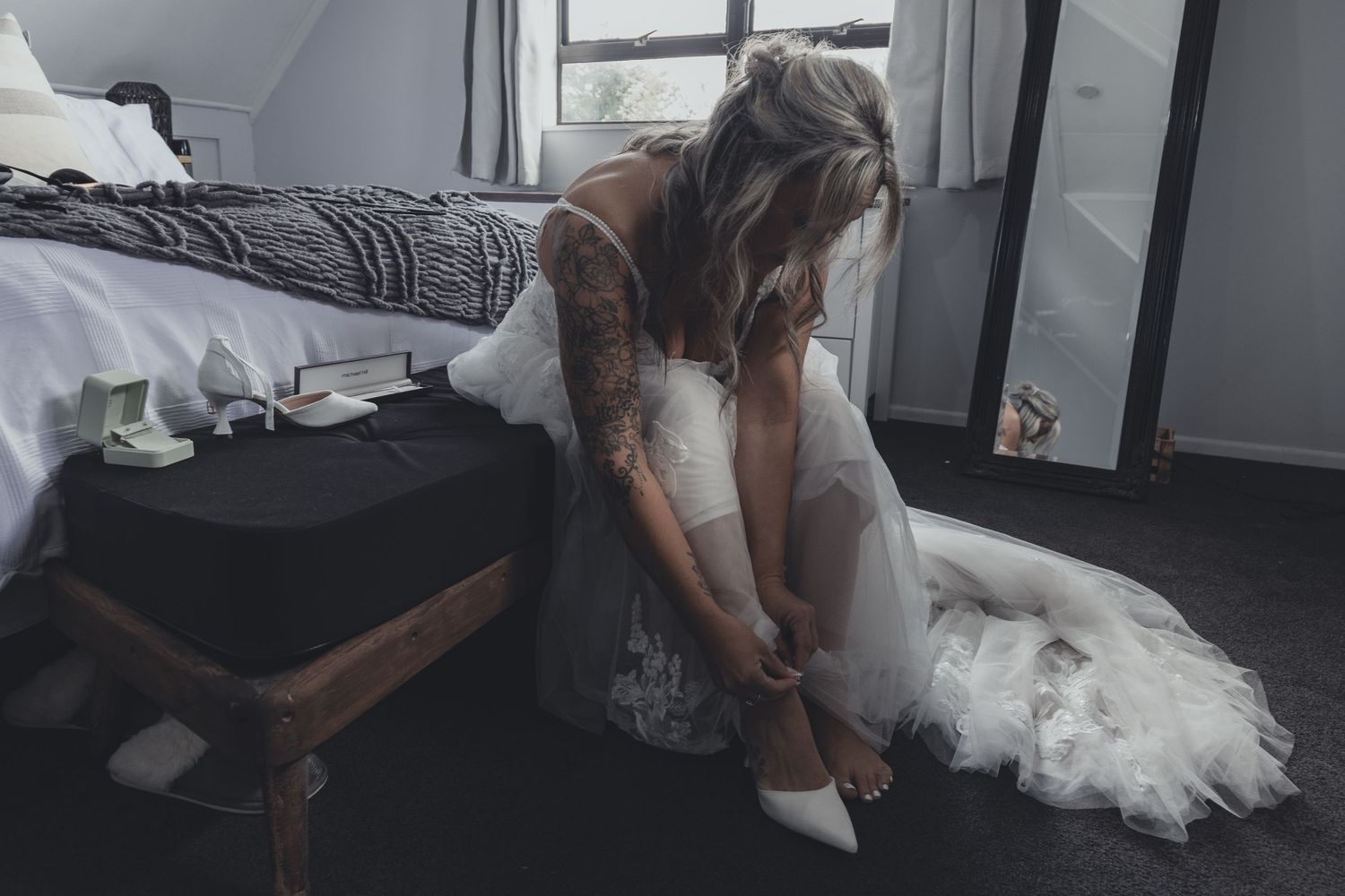 Bride in lace wedding gown putting on white heels while seated beside bed.