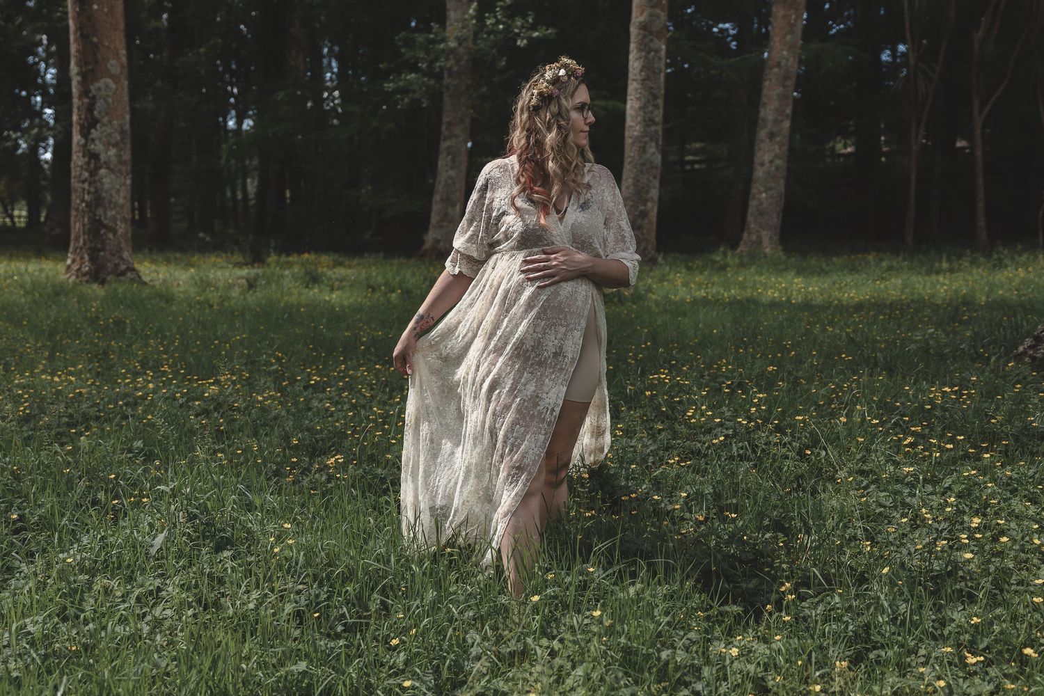 Maternity photoshoot with woman in flowing lace dress among trees in a green meadow.
