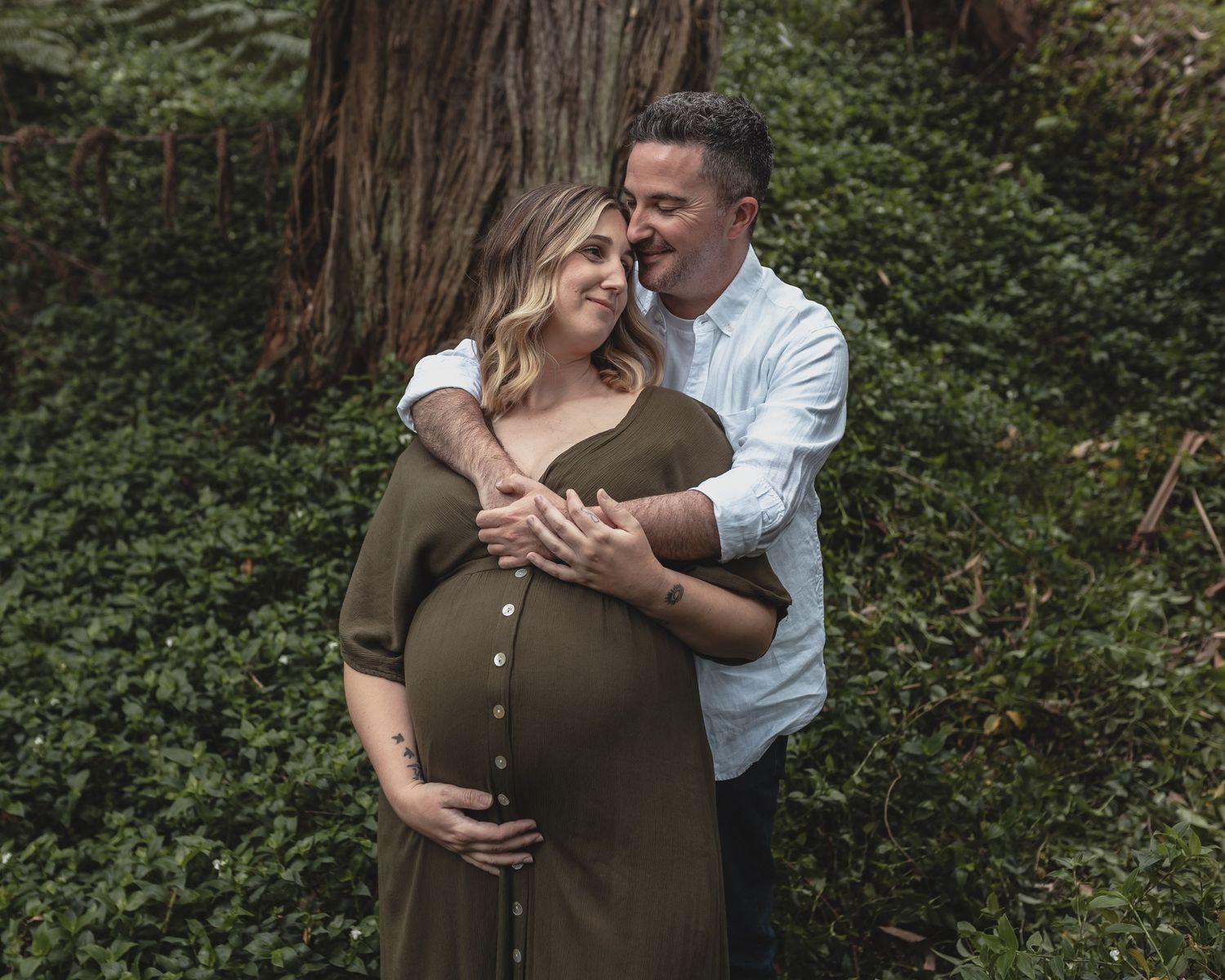 Couple sharing a tender maternity photo session outdoors among lush green trees.