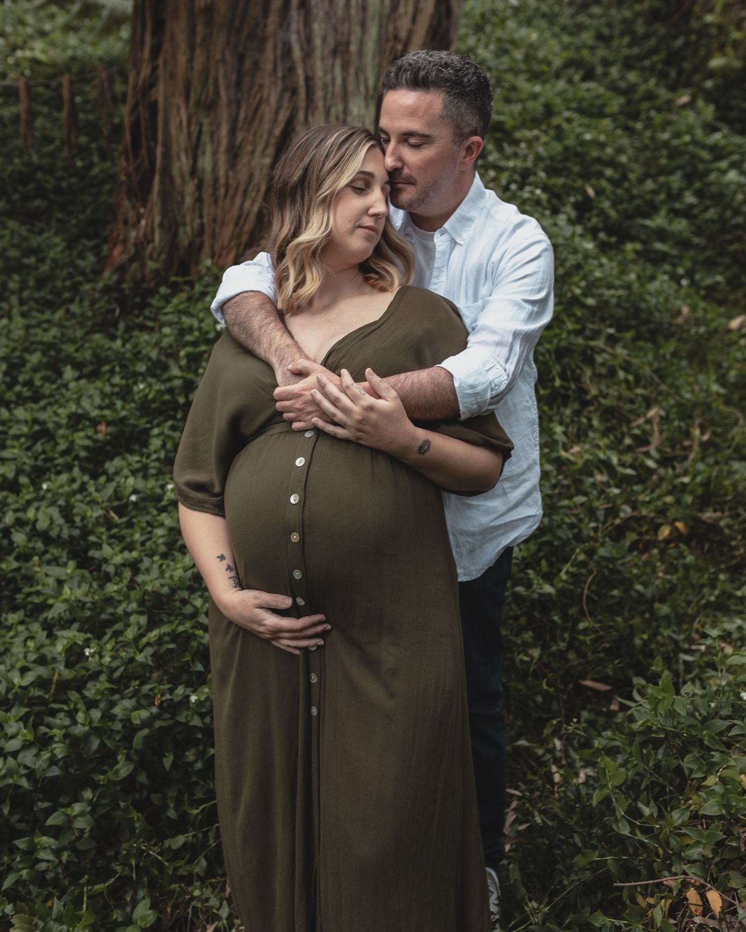 Couple sharing a tender embrace in a forest setting, man kissing pregnant woman in olive dress.