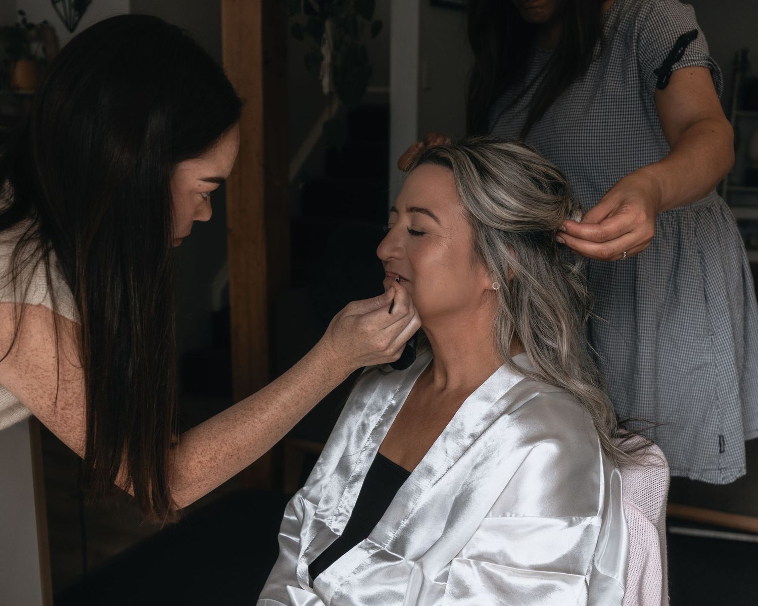 A bride in a satin robe gets makeup applied while her hair is styled.