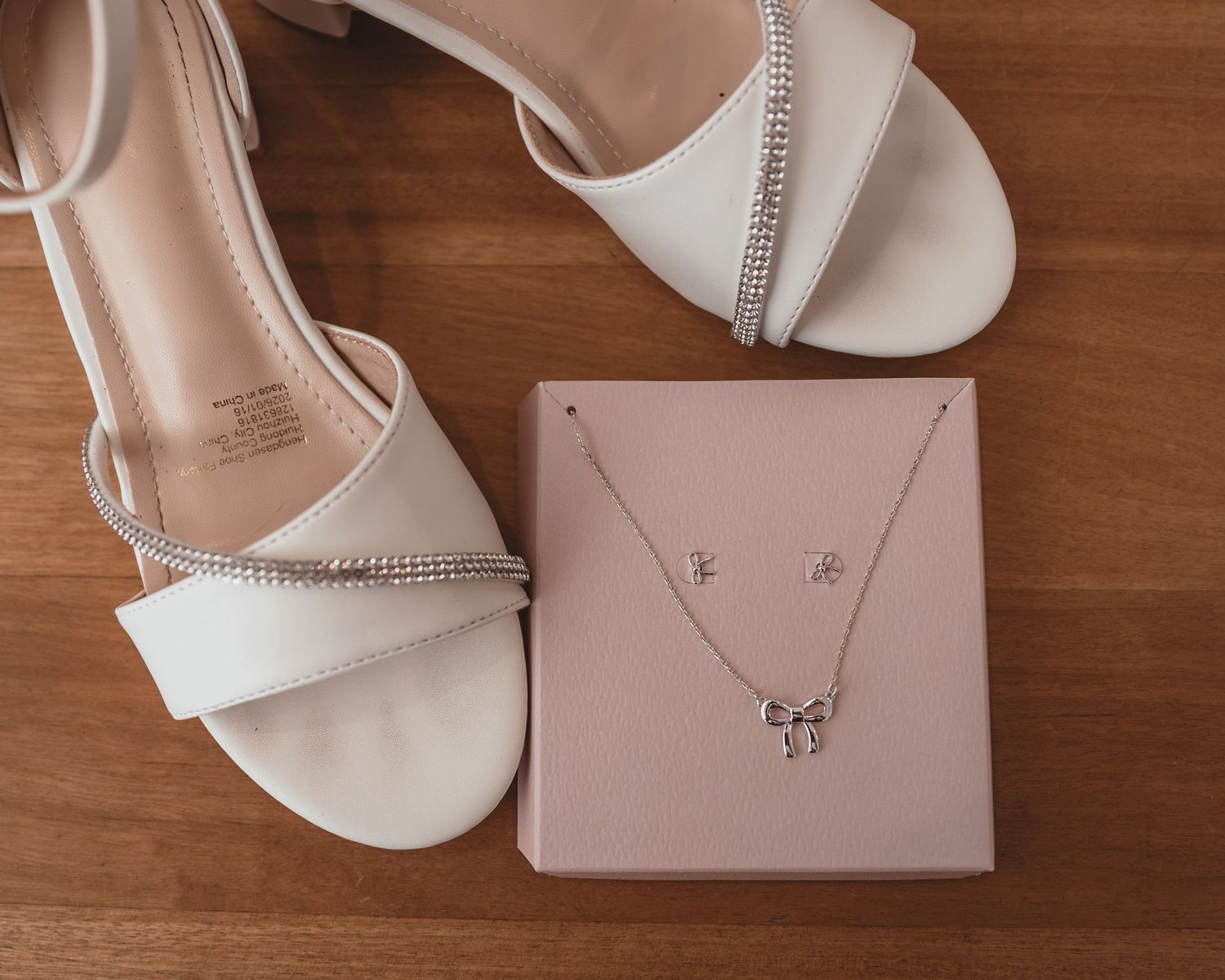 White block heel sandals with rhinestone straps beside a silver bow necklace and earrings set on a pink jewelry box.