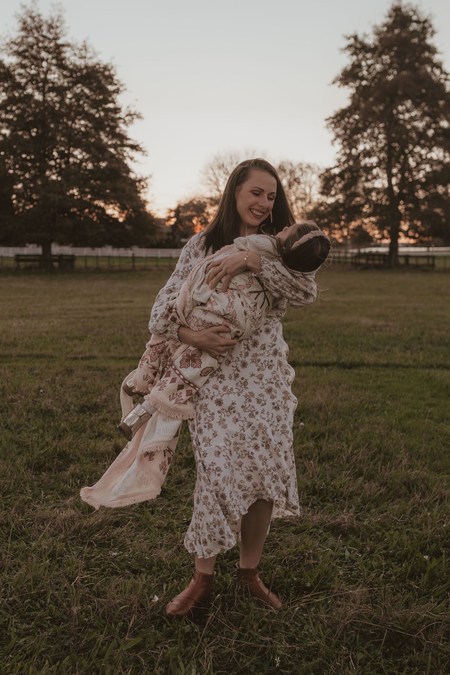 Woman in floral dress holding her baby at golden hour in a green field.