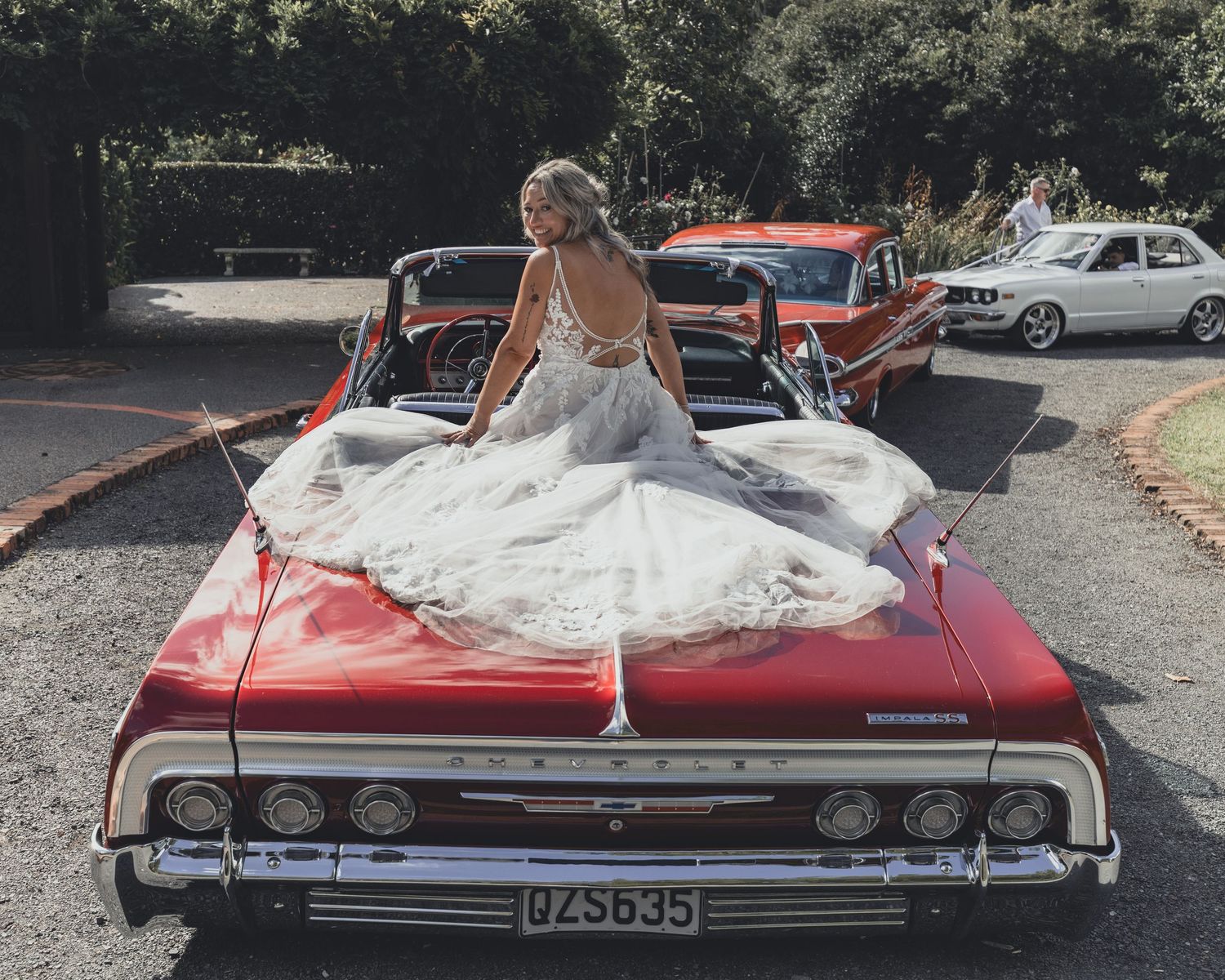 Elegant bride in backless gown sits atop vintage red classic car at wedding.