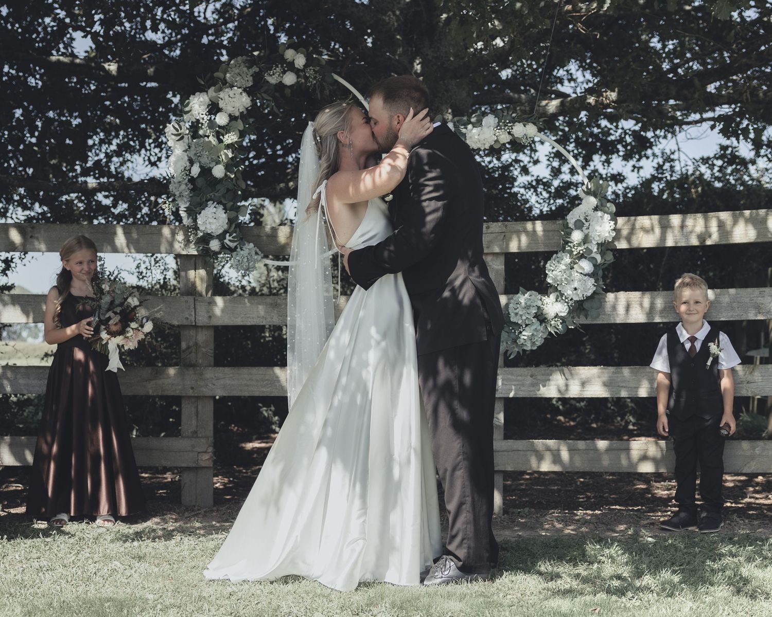 Bride and groom share first kiss at outdoor wedding ceremony near white fence with floral arch.