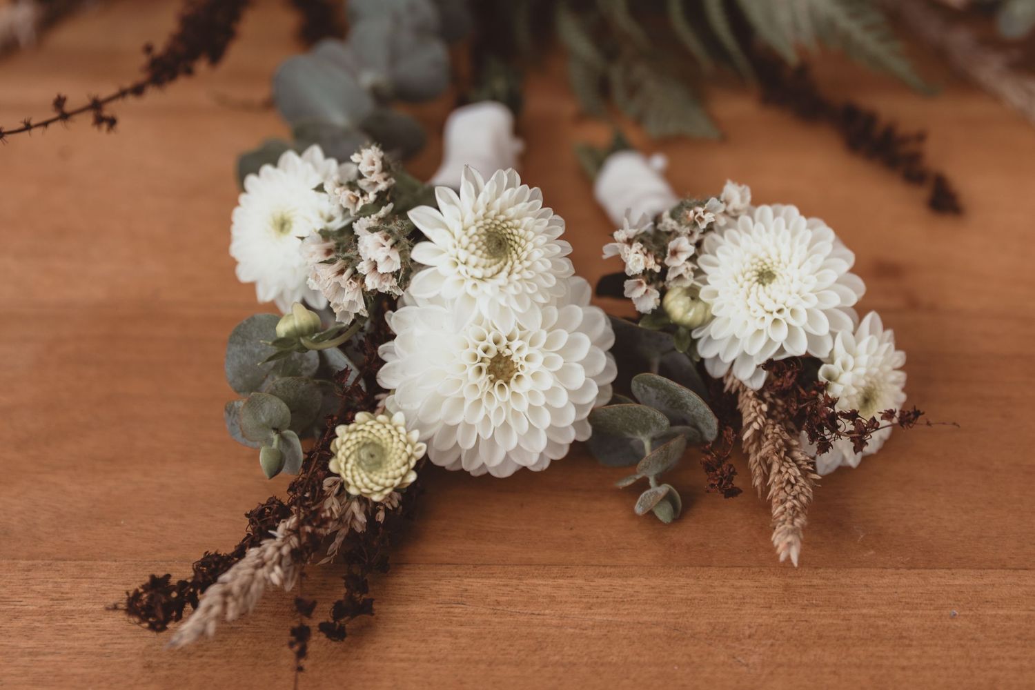 White dahlia and chrysanthemum boutonnières with eucalyptus and dried grasses on a wooden surface.