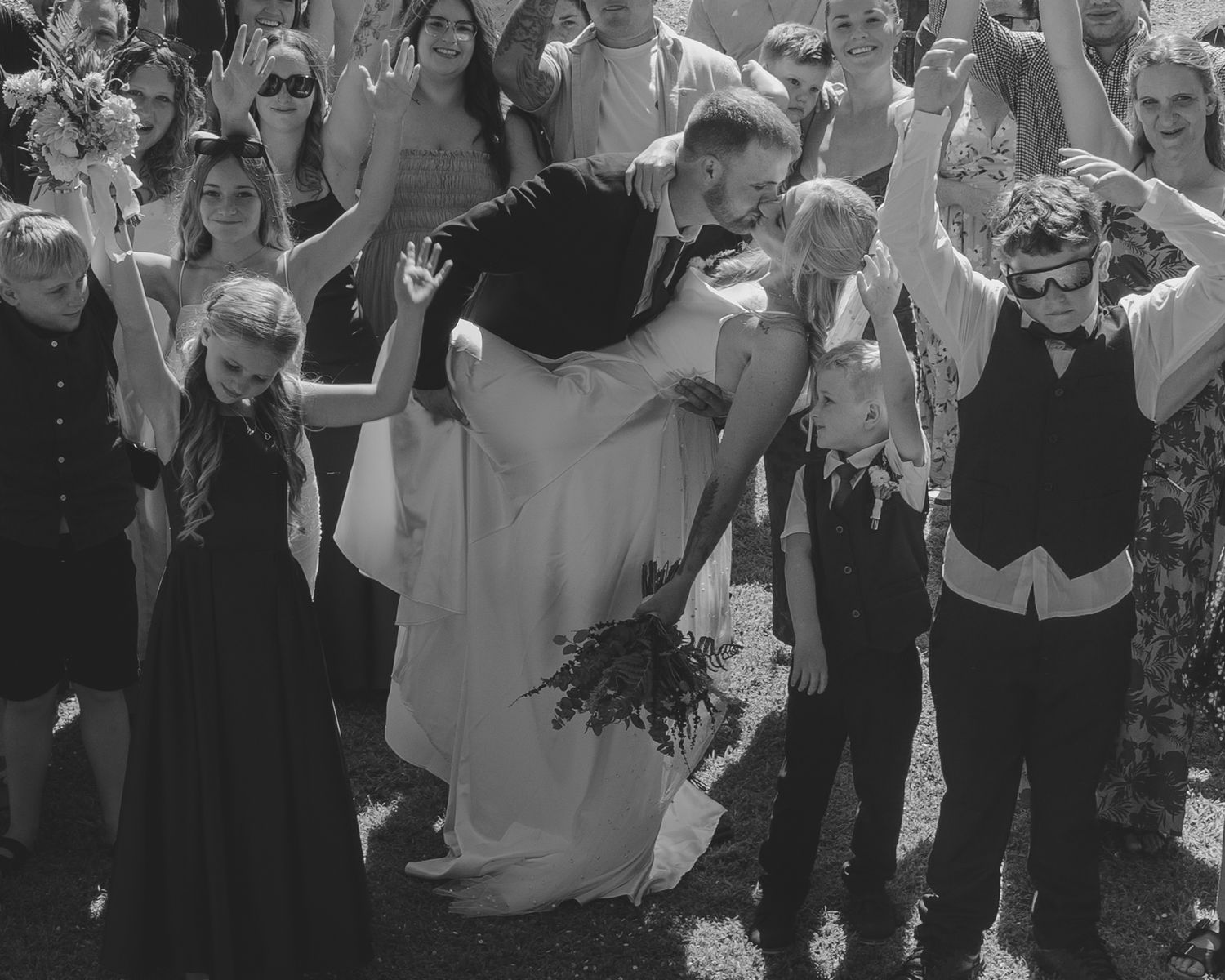 Black and white wedding photo of groom carrying bride surrounded by cheering guests outdoors.