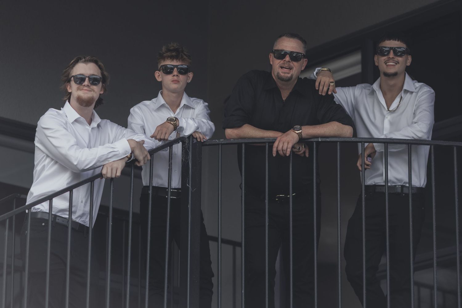 Four groomsmen in white shirts and sunglasses posing together on outdoor balcony railing.