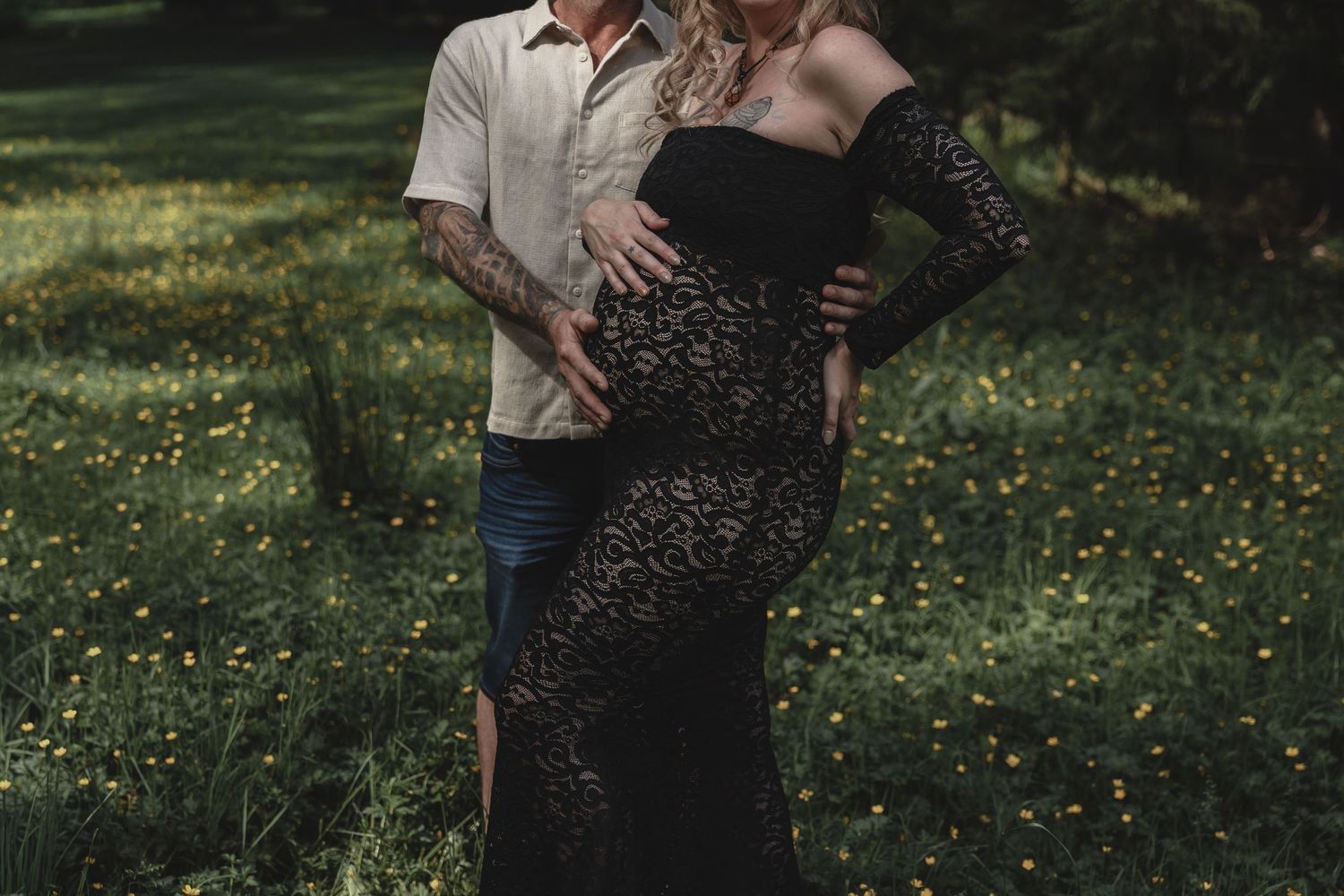 Couple embracing during maternity photoshoot in a meadow, man with tattooed arms.