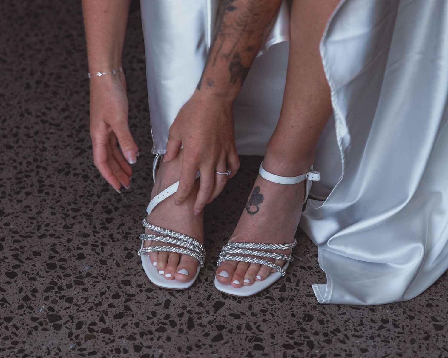 Bride adjusting white strappy heeled sandals with rhinestone details, wearing a white wedding dress, tattoo visible on leg.