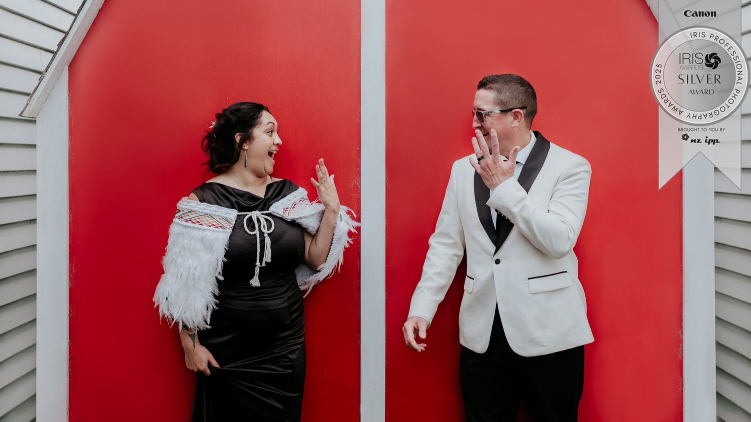 Two people pose playfully in front of red doors, one in a black fringe dress and one in a white tuxedo jacket, photography by Charly Baty.