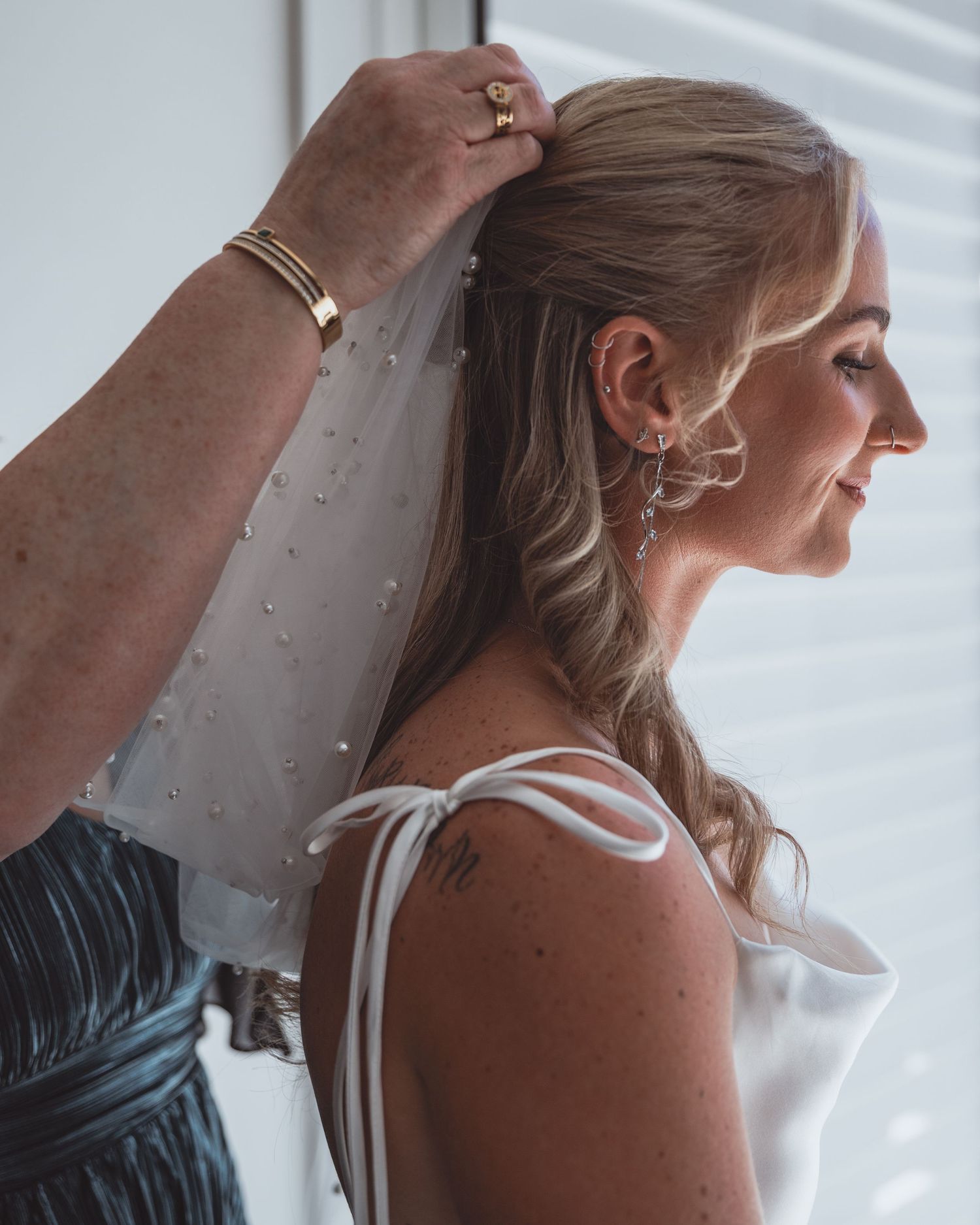 A bride smiles as her veil is pinned into her blonde hair, wearing a white gown with elegant drop earrings.