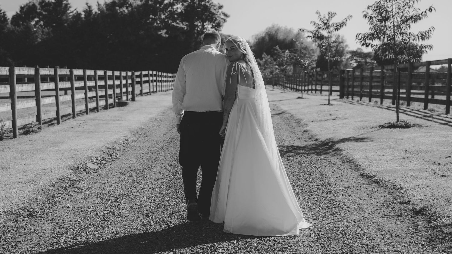 Black and white photo of a bride and groom walking hand in hand down a gravel path lined with wooden fences.