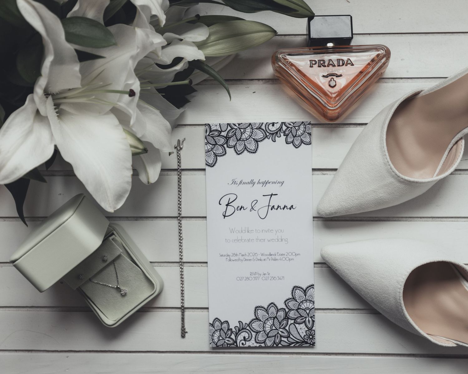 A wedding invitation with lace design displayed beside heels, jewelry, and a Prada perfume.