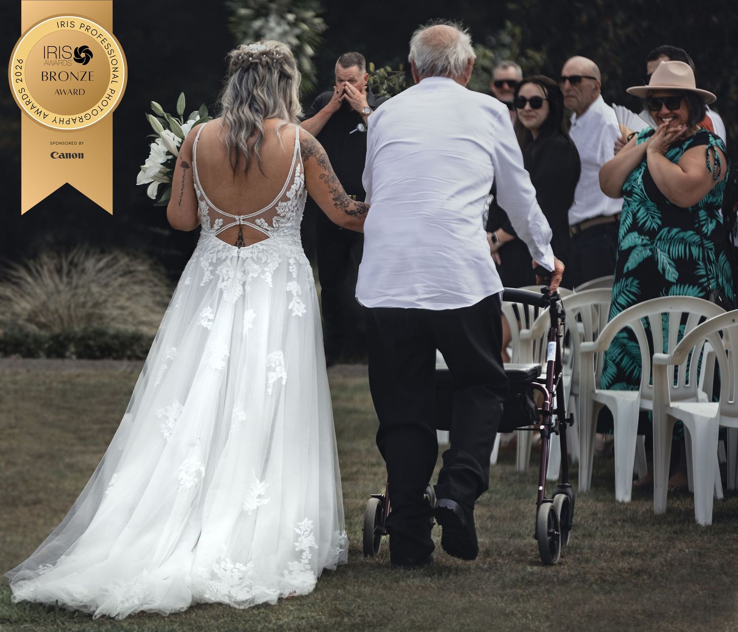 Bride in backless white gown walks dad with walker at outdoor wedding ceremony, award-winning photo, by Charly Baty.