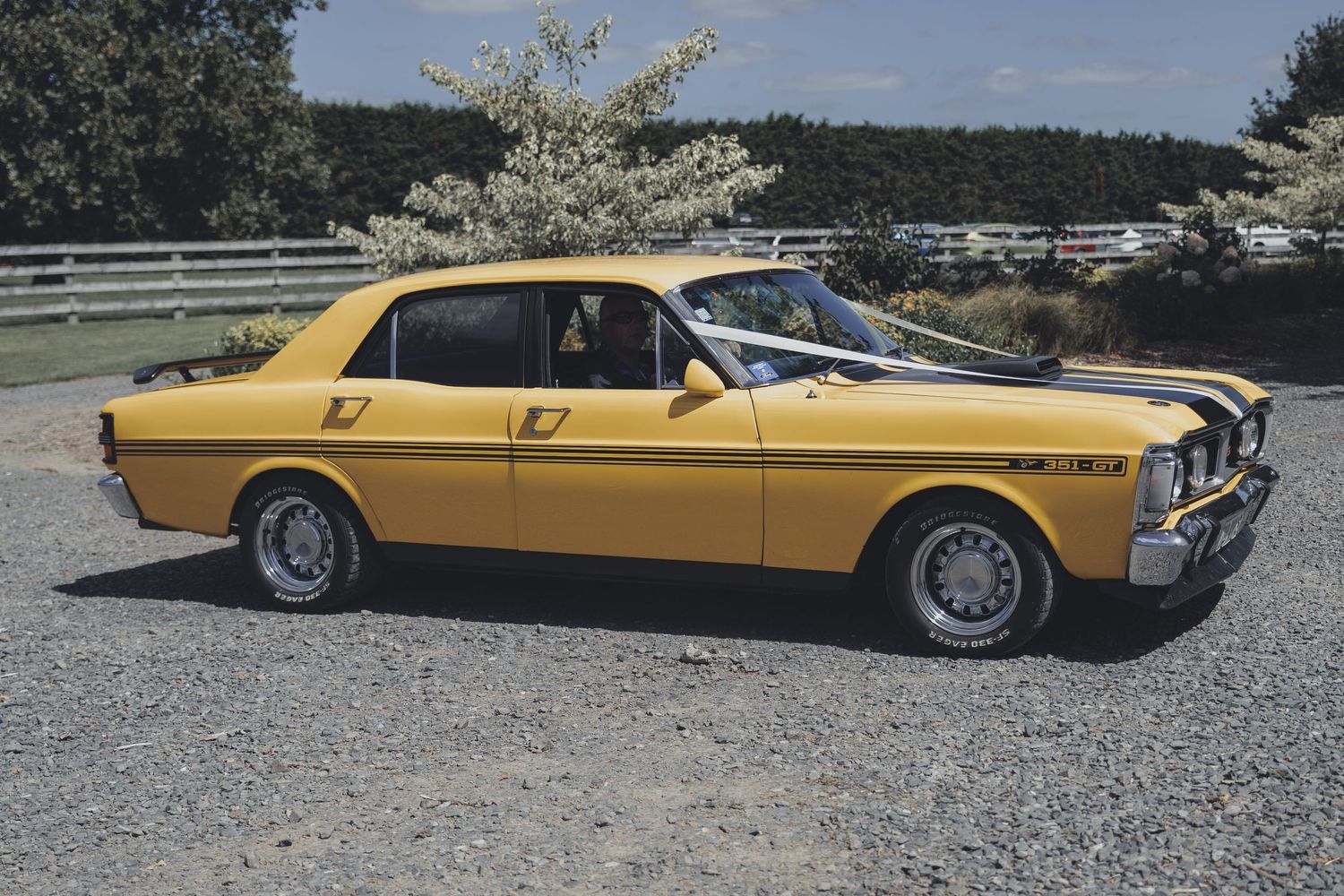 Classic yellow Ford Cortina muscle car parked on gravel driveway with trees in background.