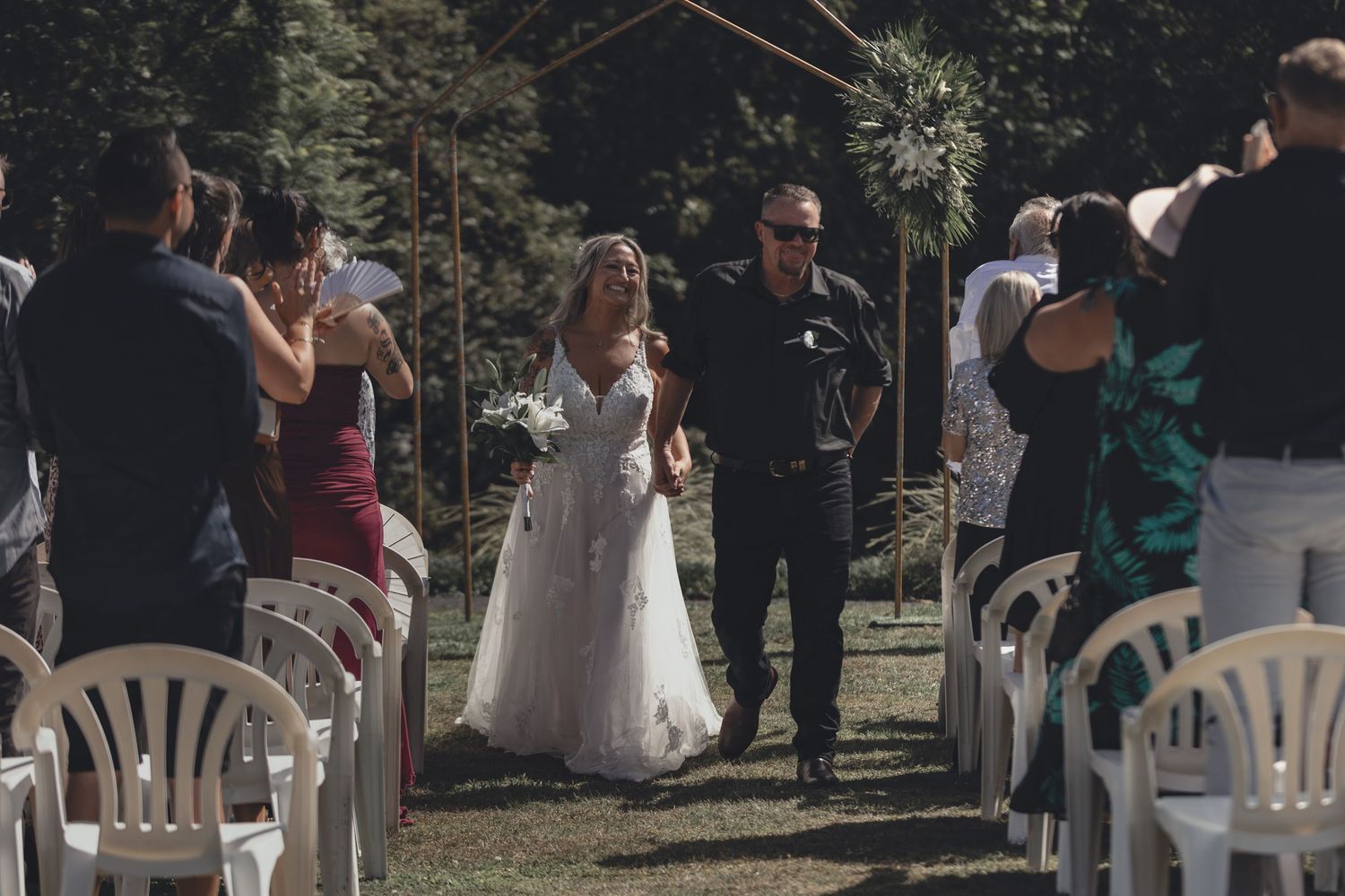 Bride walks down outdoor wedding aisle with escort among seated guests in white chairs.