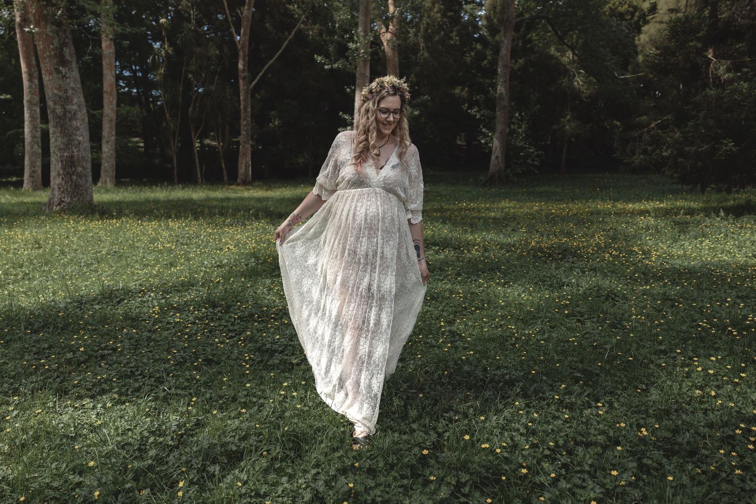 Pregnant woman in elegant white lace gown posing in a lush forest clearing.