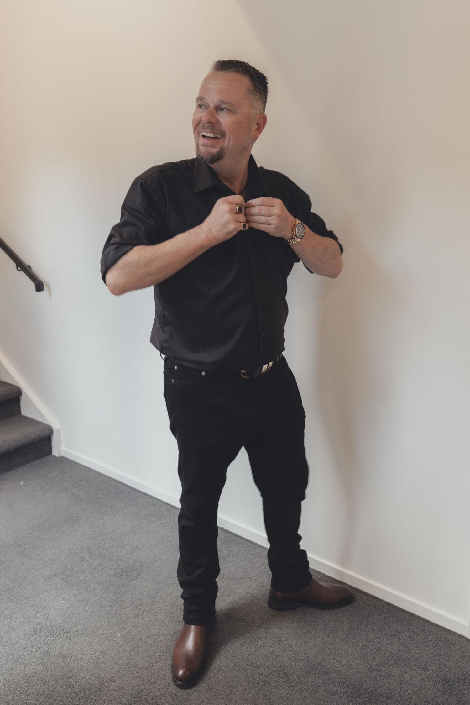 Groom in black shirt and pants buttoning collar while smiling on staircase landing.