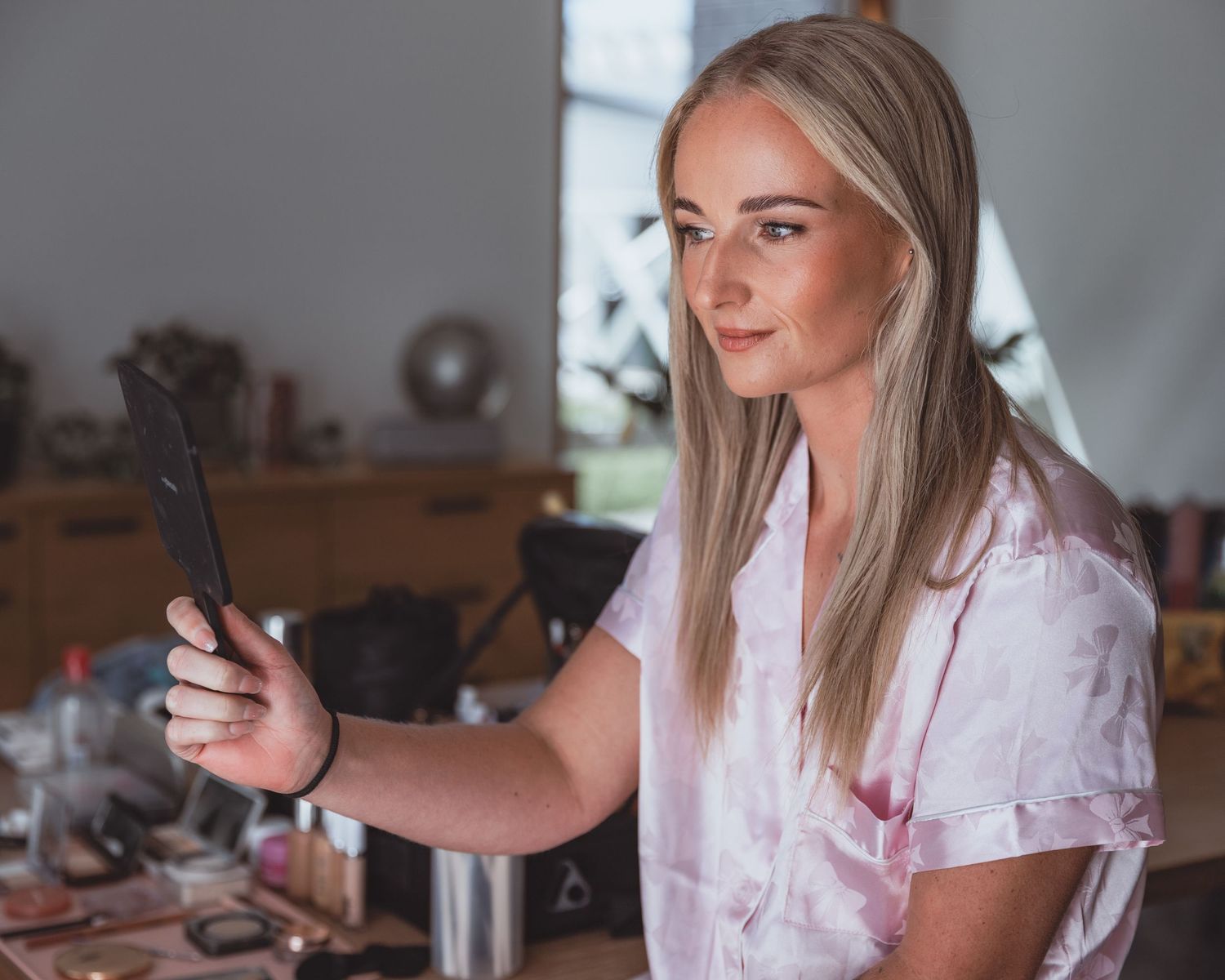 Young blonde woman in pink satin top taking a selfie with smartphone near a makeup vanity table.