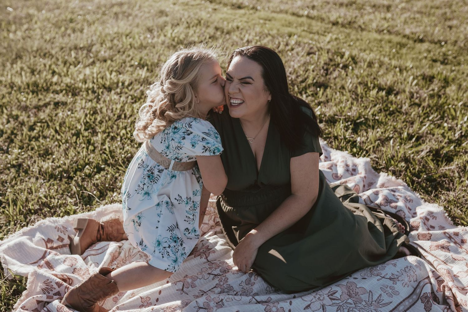 Young girl whispering to her smiling mother while sitting on a blanket in a grassy field.