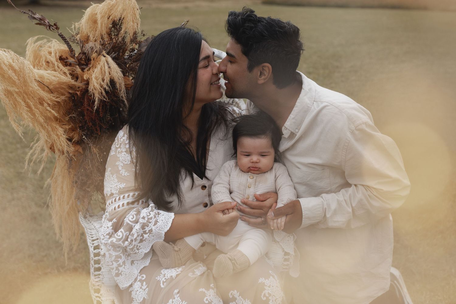 Couple sharing a kiss while holding their newborn baby during an outdoor family photoshoot.